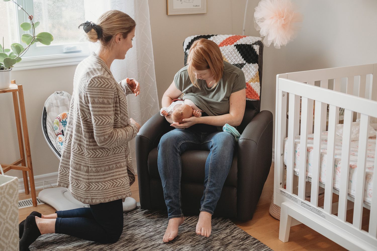 Two women in a nursery, one kneeling beside a chair where the other sits breastfeeding a newborn baby near a white crib.