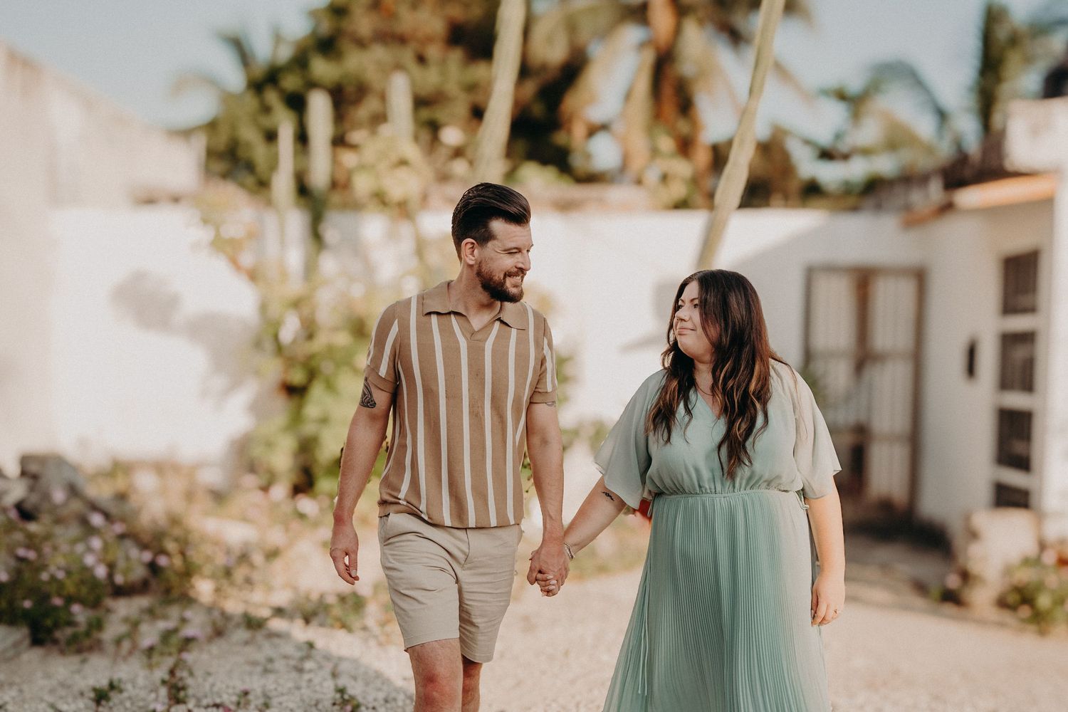 A couple holds hands and smiles while walking outdoors near palm trees, the woman in a mint dress and man in a striped shirt.