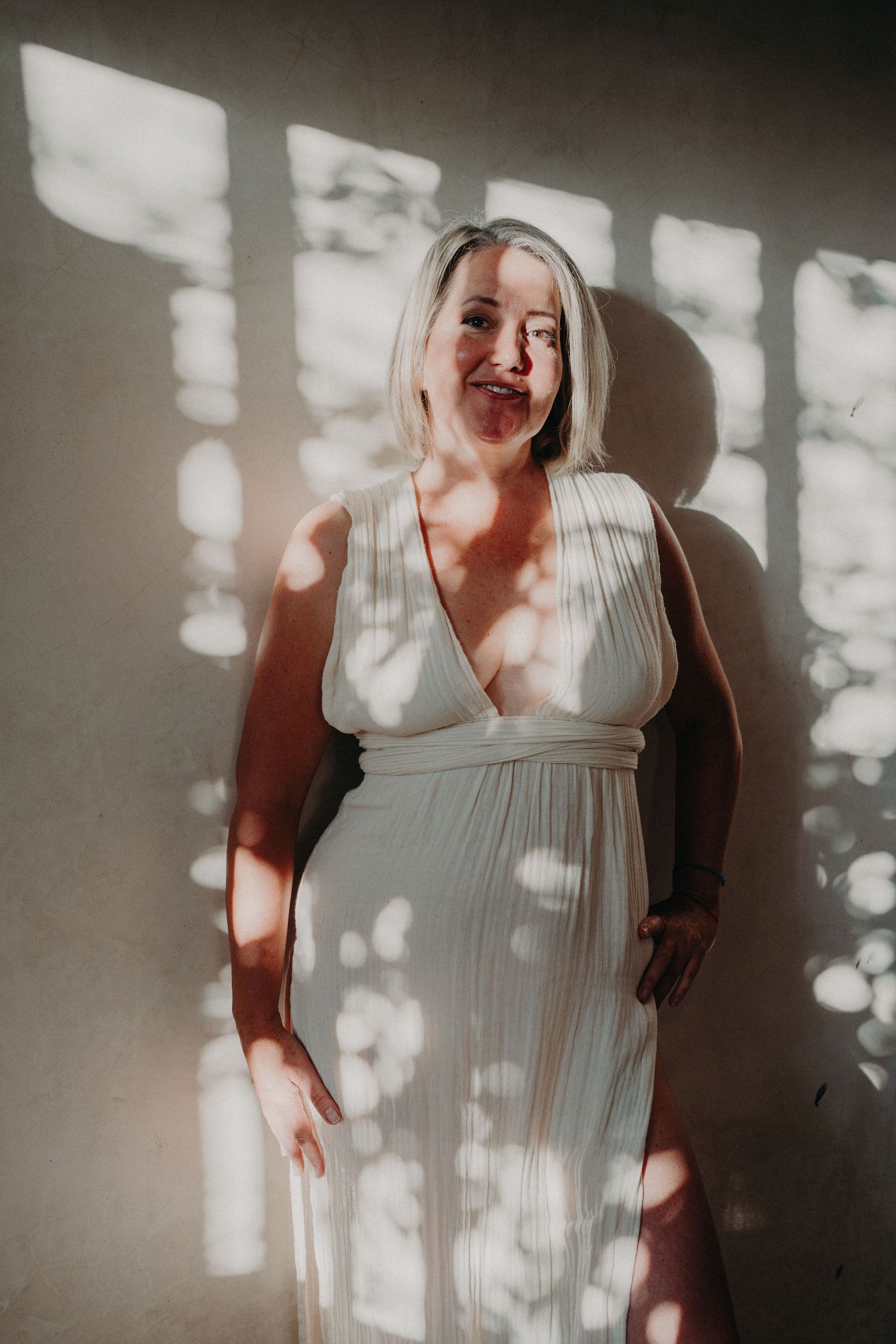 Pregnant woman in white dress posing against sunlit wall with dappled light shadows.