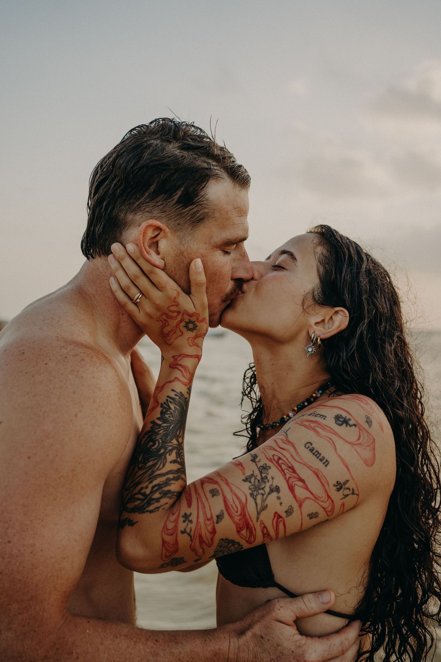 Shirtless man and woman with wet hair and colorful arm tattoos share a romantic kiss while standing in the ocean at sunset.