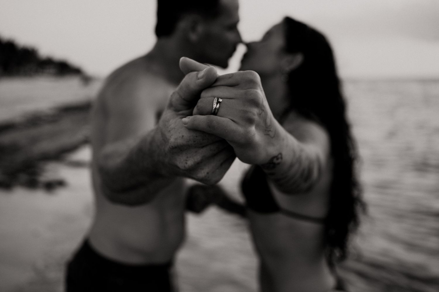 Black and white photo of a barefoot couple in swimwear sharing a kiss on a beach, their clasped tattooed hands and ring in sharp focus.