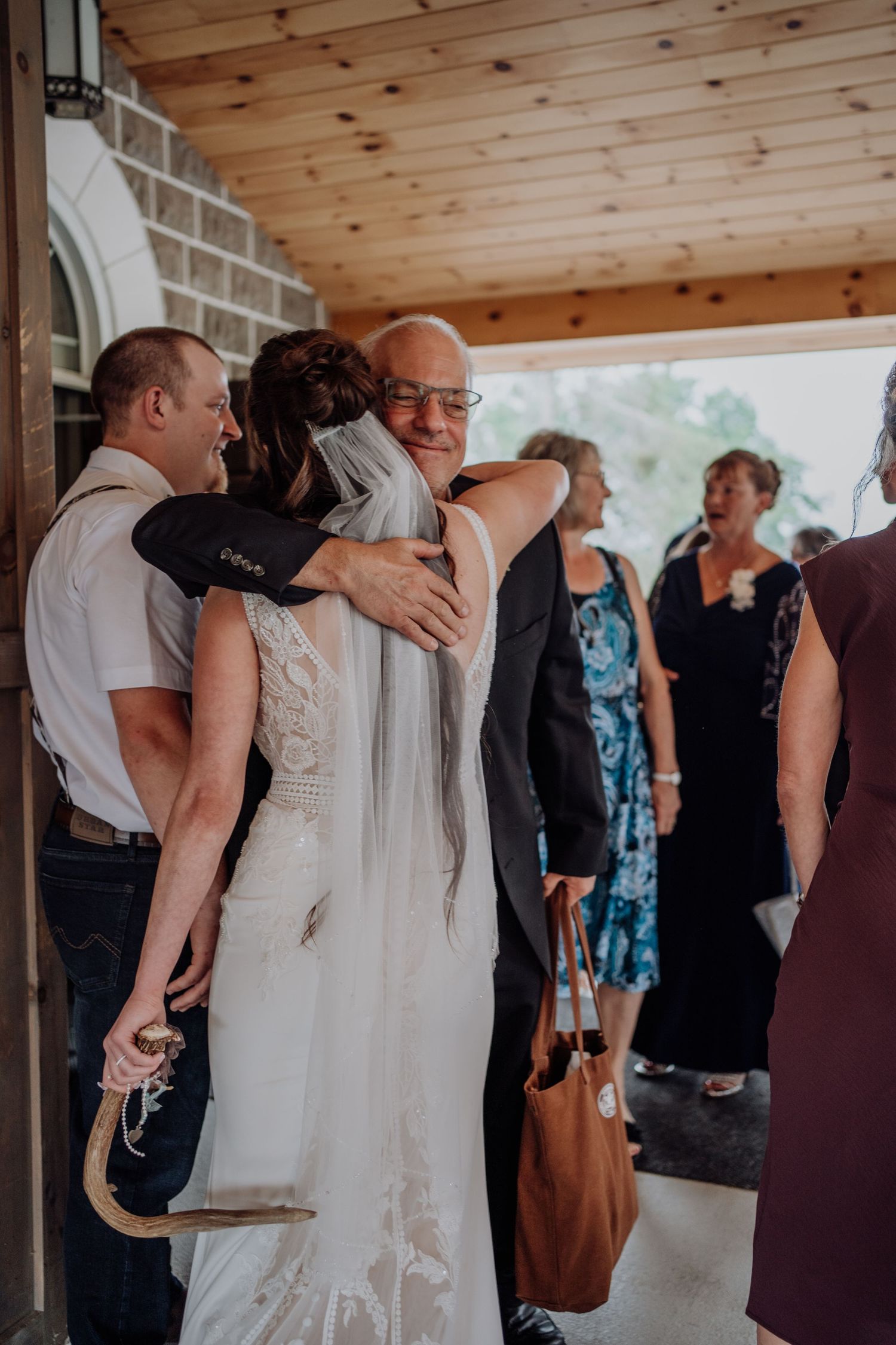Bride in white wedding gown embracing older gentleman guest under wooden porch after ceremony.
