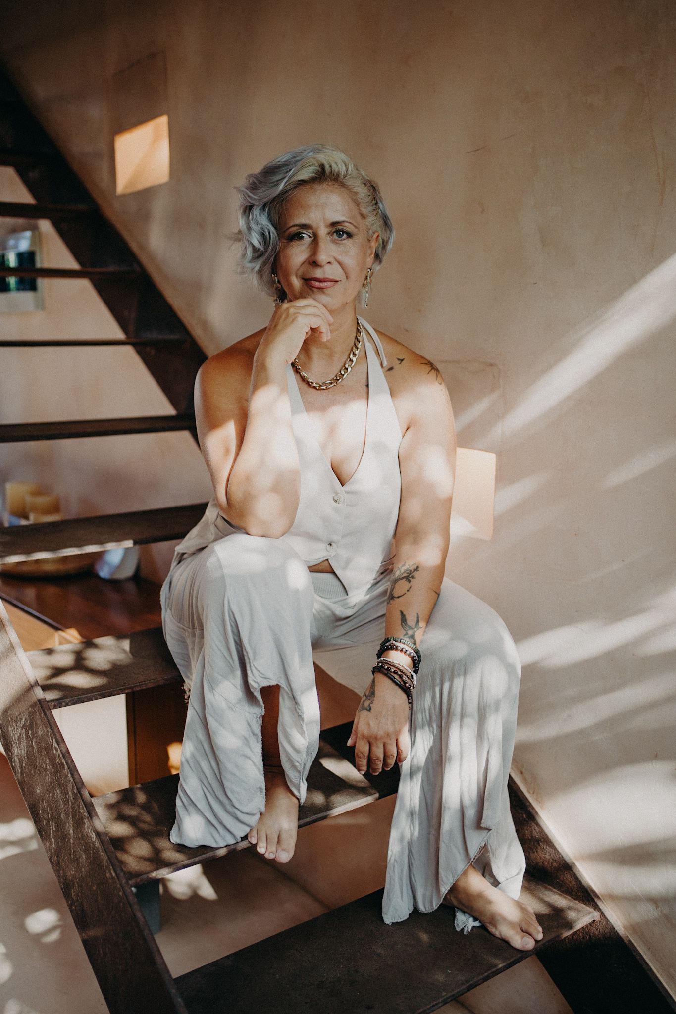 Mature woman with blue hair sitting on wooden stairs wearing a white jumpsuit and layered jewelry.