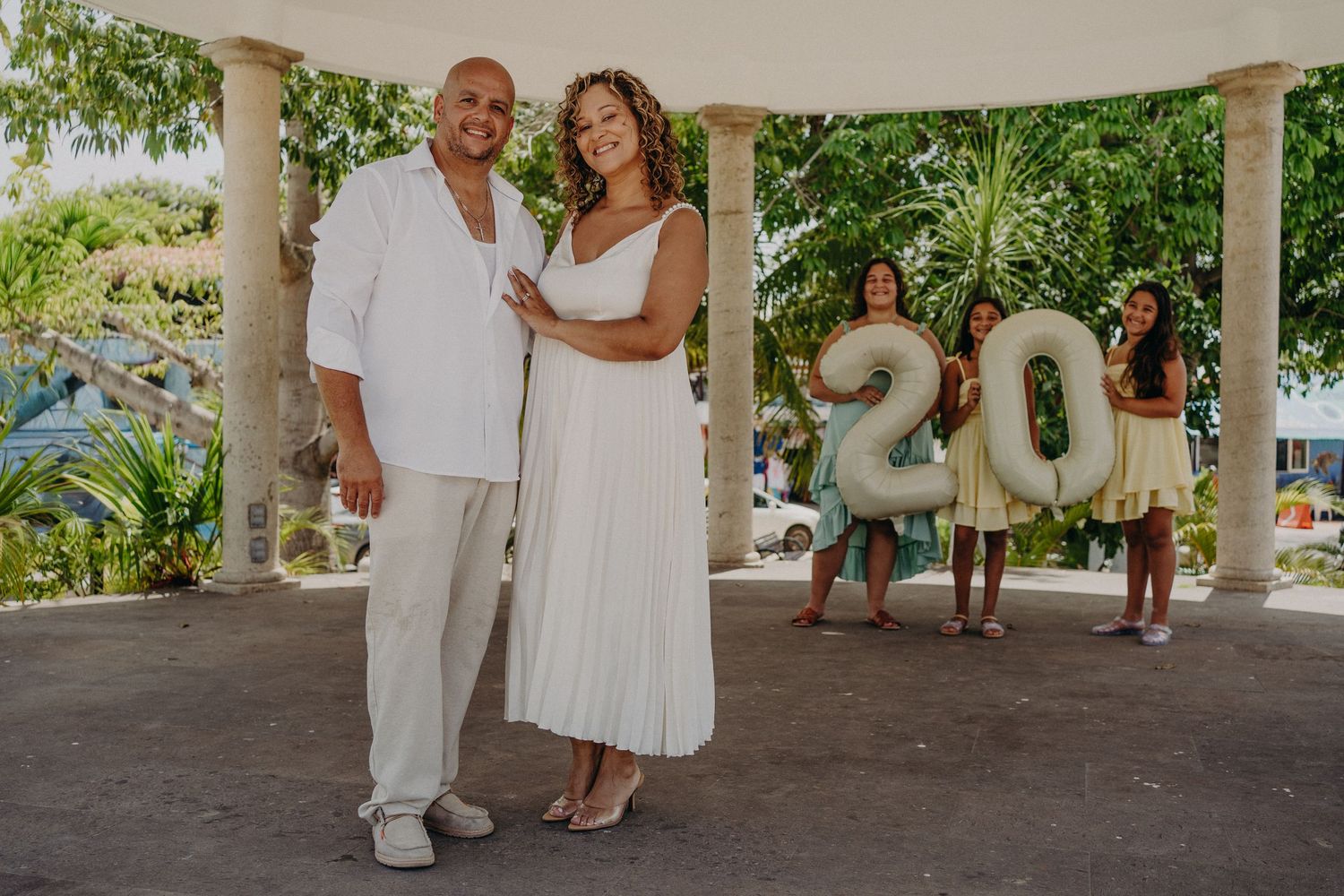 Couple in white outfits celebrating 20th anniversary under gazebo with two women holding balloon numbers in background.