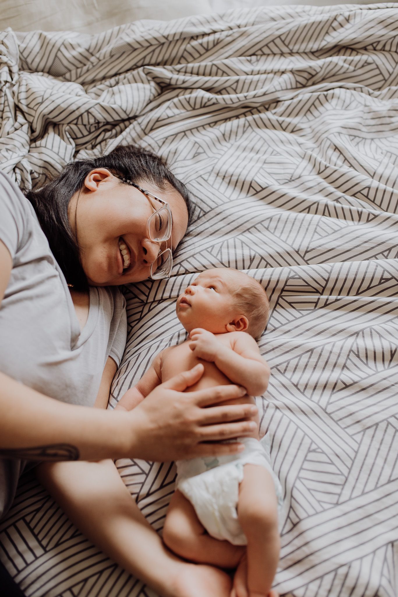 Smiling mother lying on striped bedding next to her newborn baby in a diaper, sharing a tender moment.