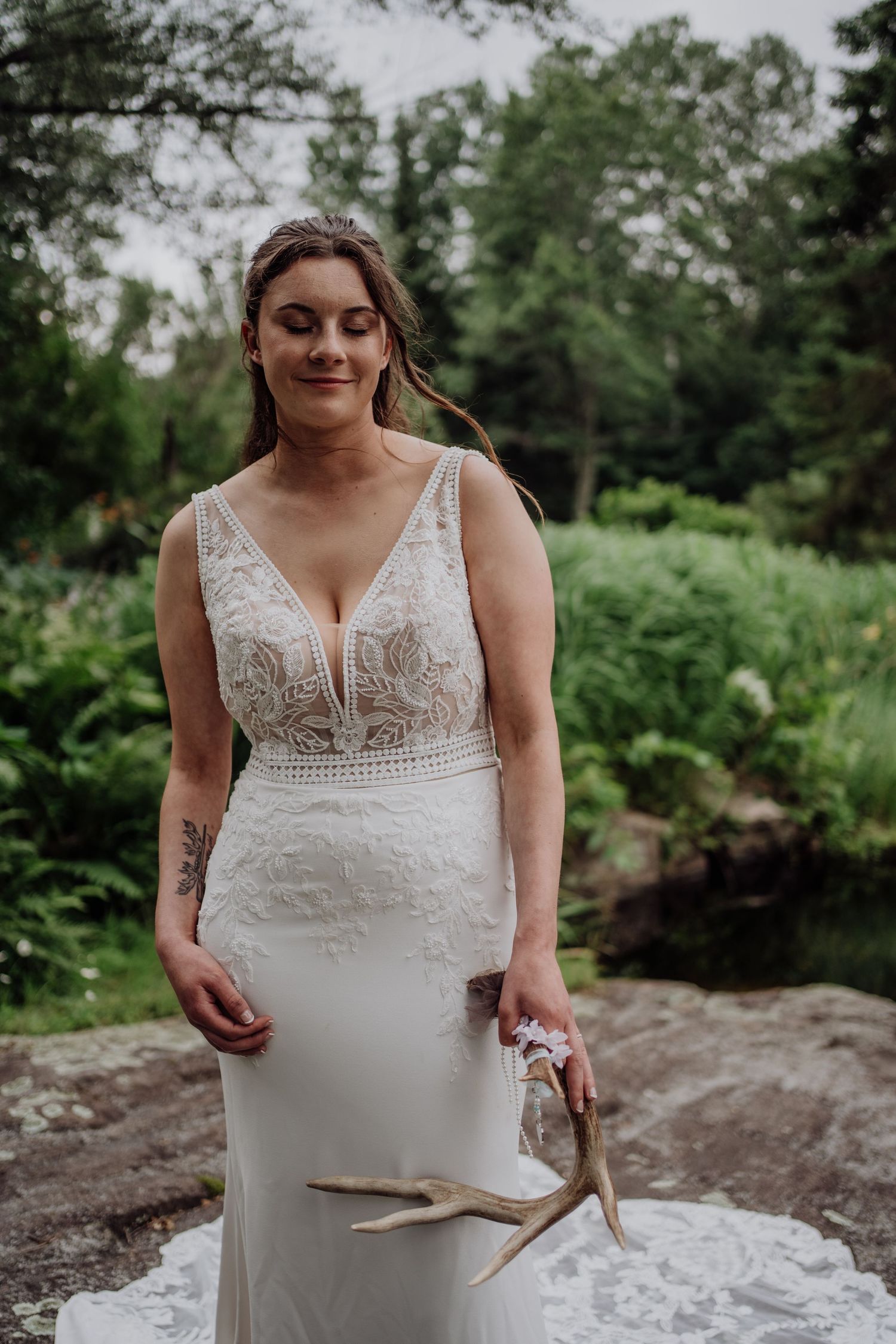 Bride in deep V-neck lace wedding dress holding deer antlers outdoors with green forest background.