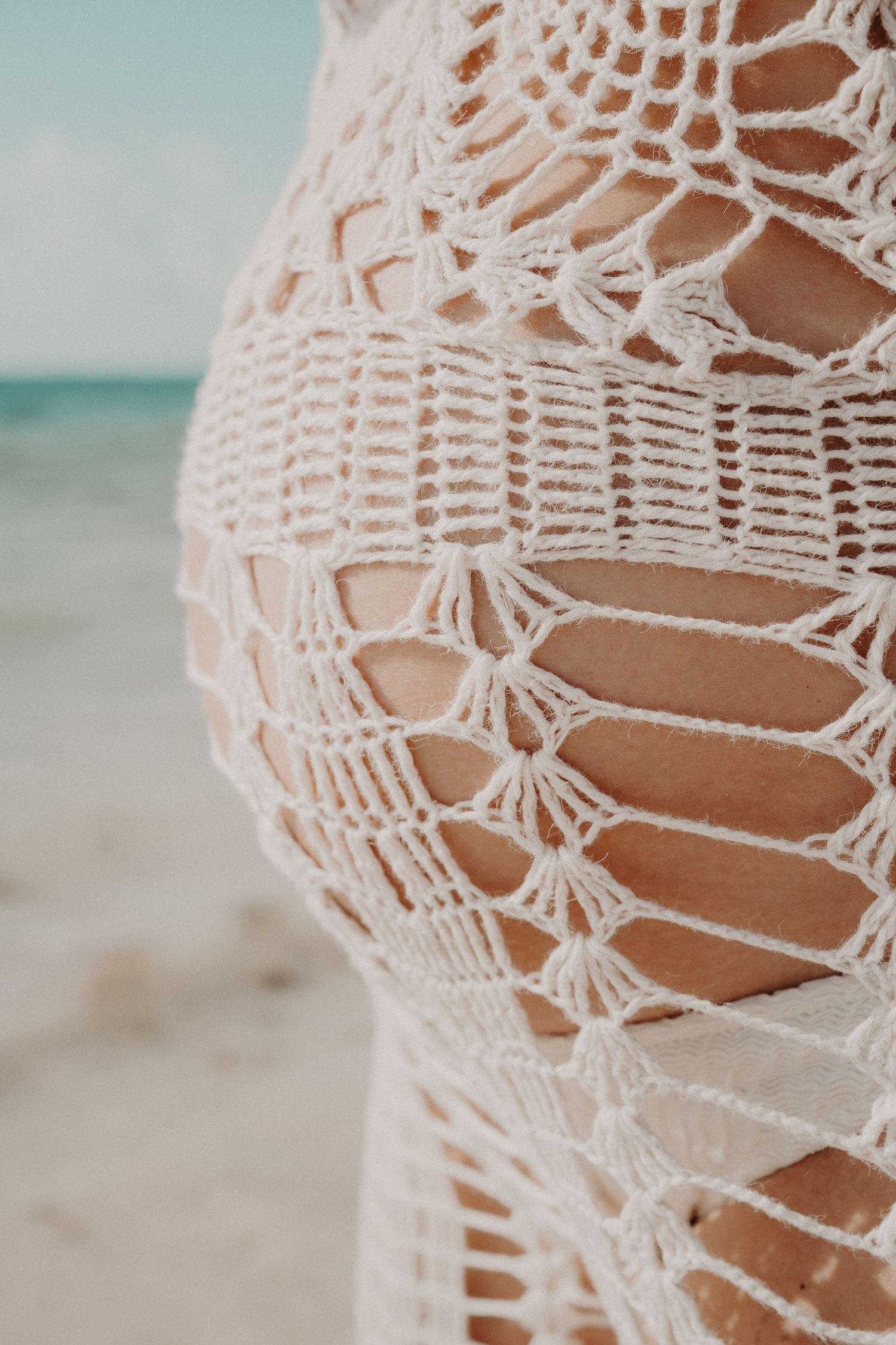 Close-up of a white crochet beach cover-up with an intricate lace pattern worn on a sandy shore with turquoise ocean in the background.