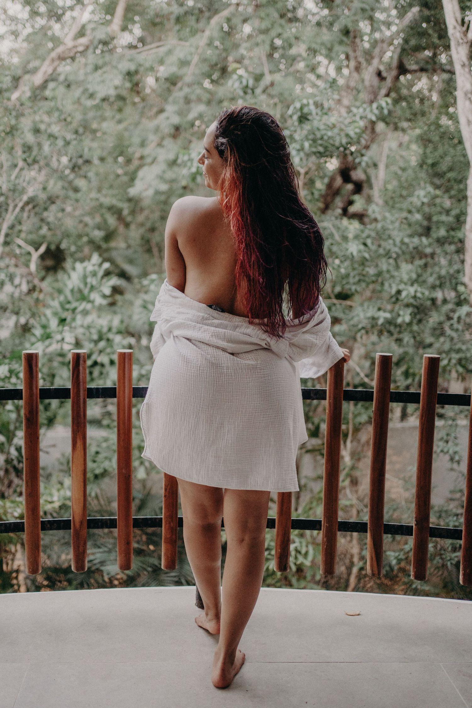 Woman with long red-tipped hair stands on a balcony wrapped in a white shirt, overlooking a lush green forest.