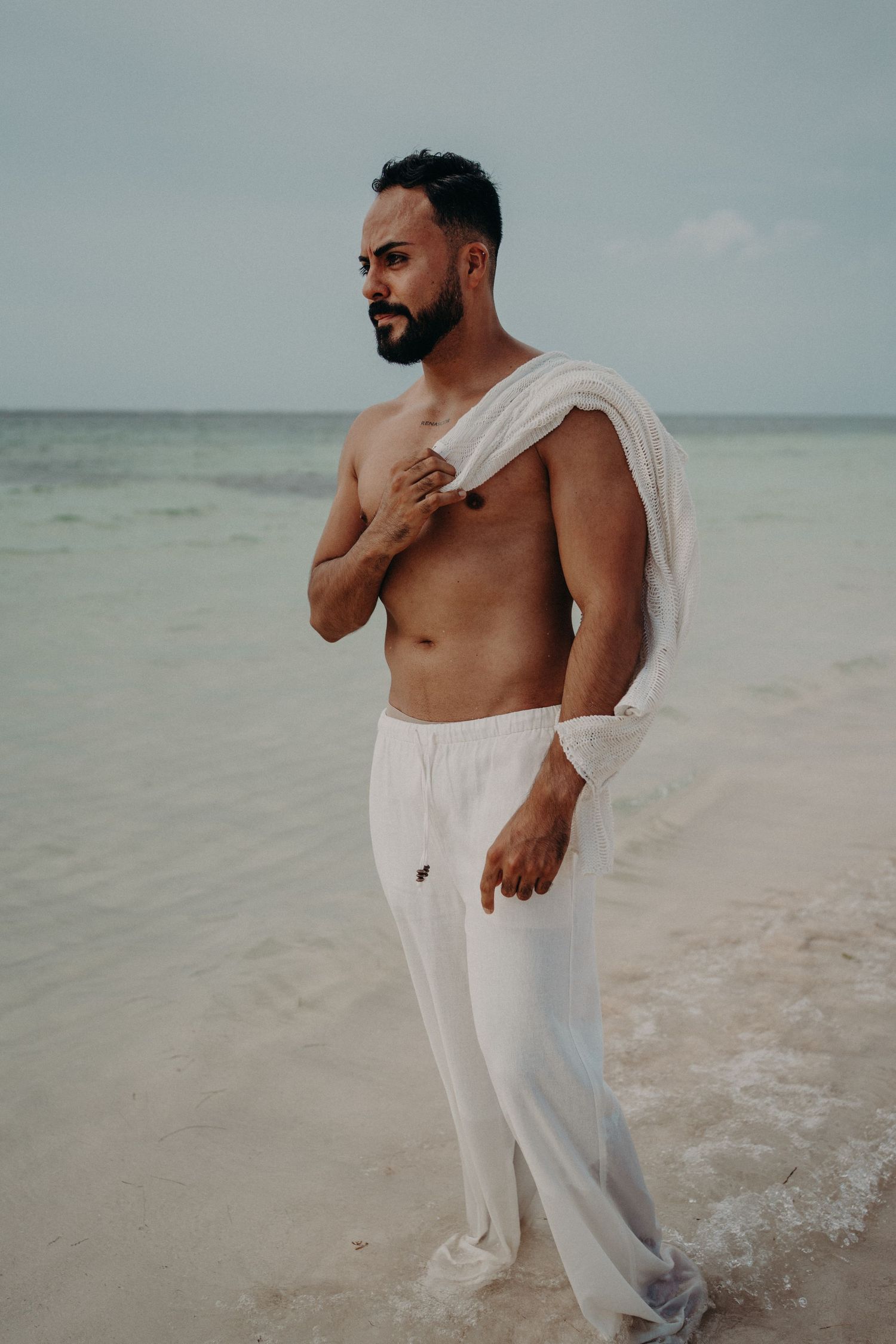 Shirtless man with beard wearing white pants standing on a tropical beach holding his shirt over his shoulder.