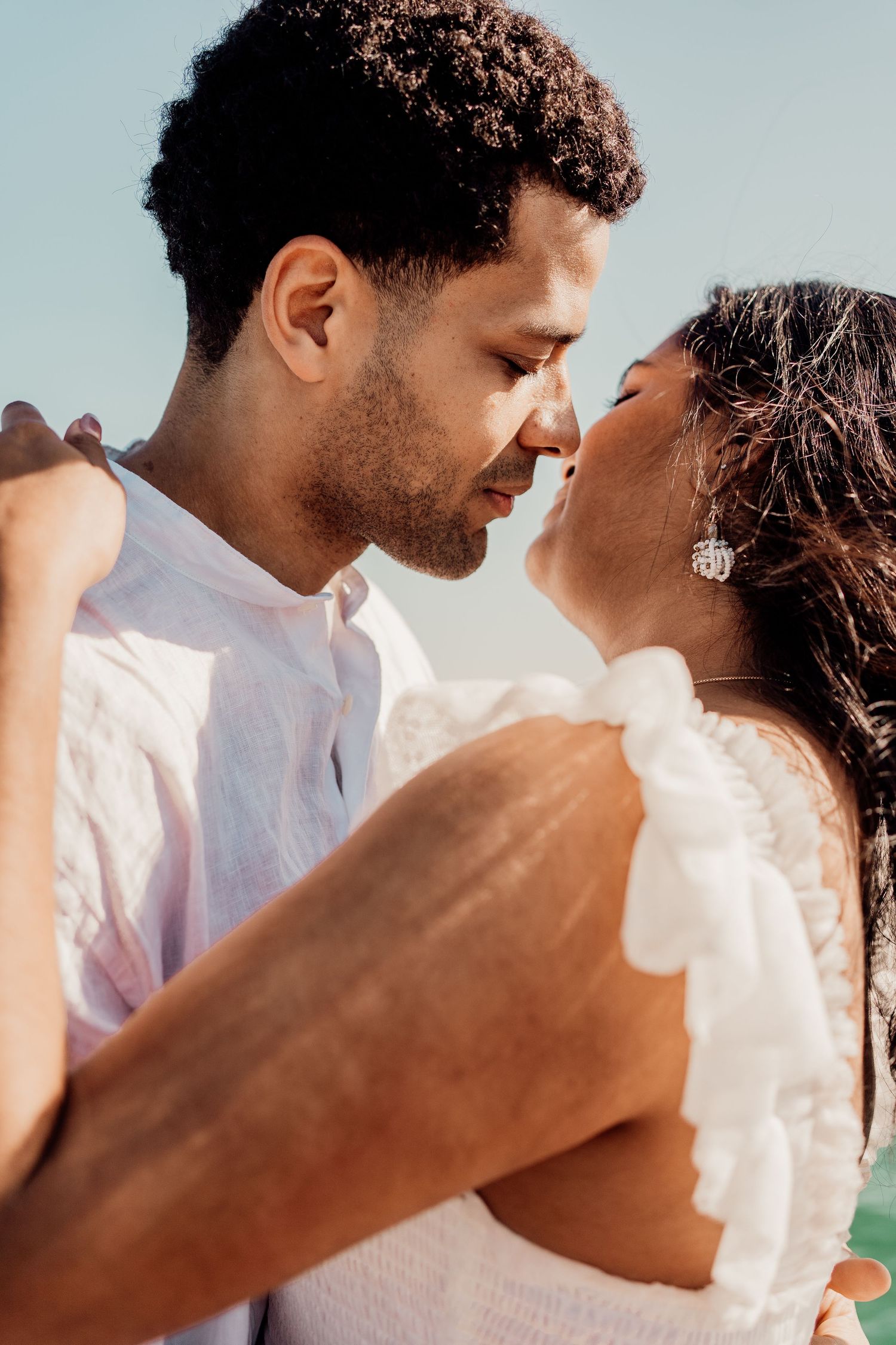 A romantic couple in white outfits share an intimate moment, nearly kissing outdoors under a clear blue sky.
