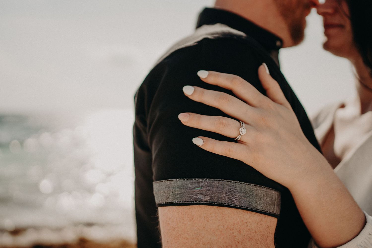 Close-up of a woman's hand with an engagement ring resting on a man's shoulder near the ocean.