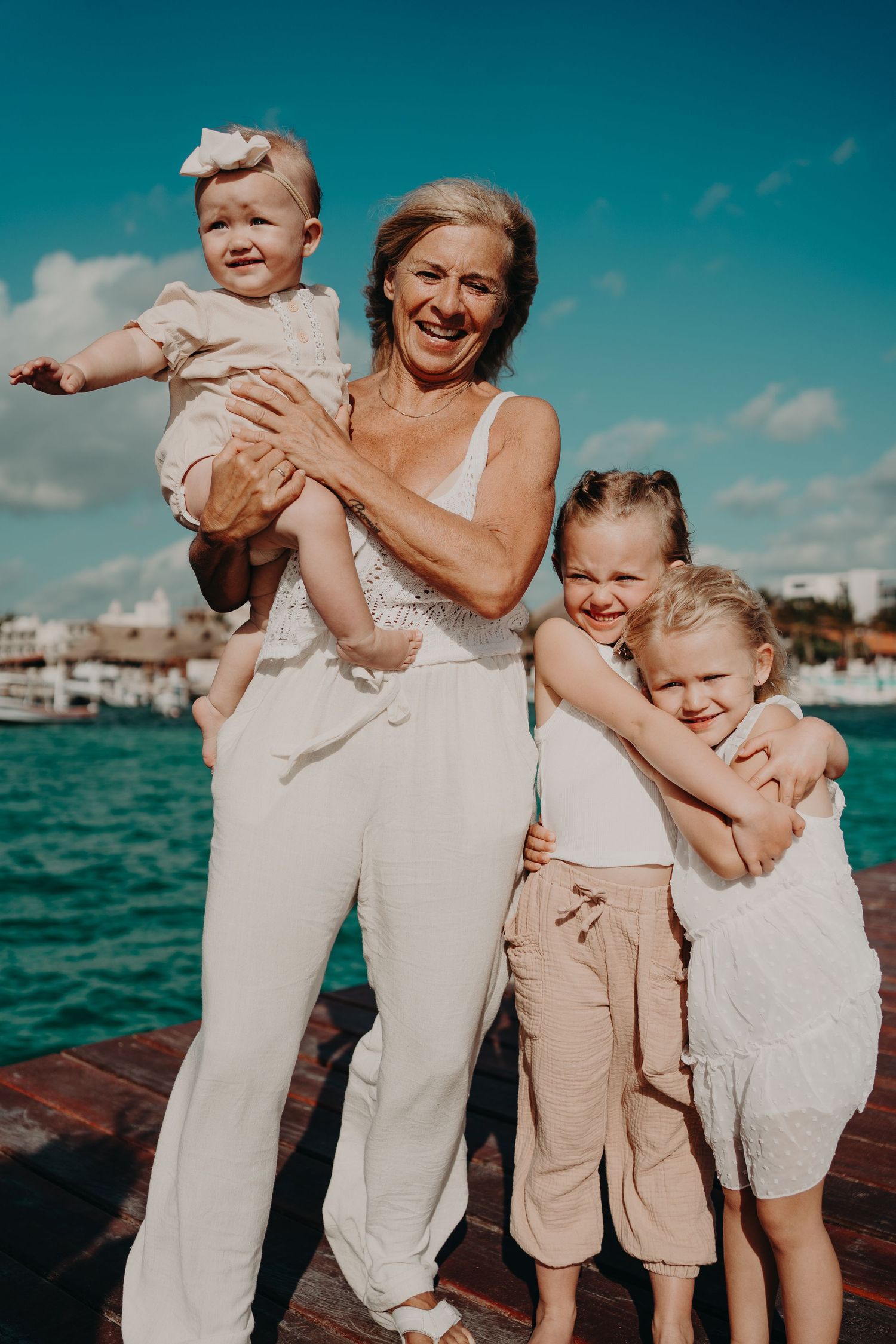 Smiling grandmother holds baby girl while two young granddaughters hug her on a sunny dock by turquoise water.
