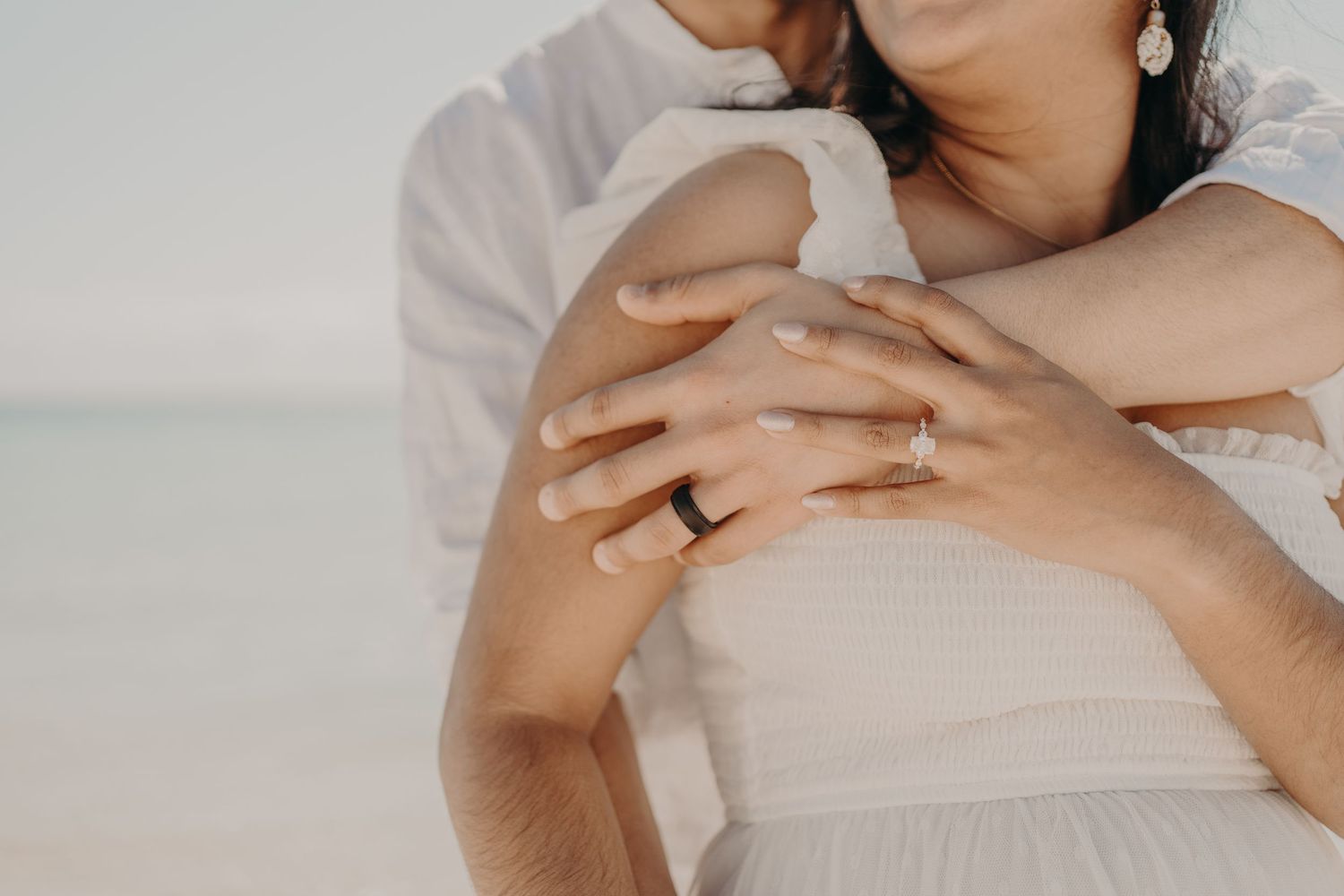 Couple embracing on beach, woman wearing engagement ring, romantic seaside wedding photo.