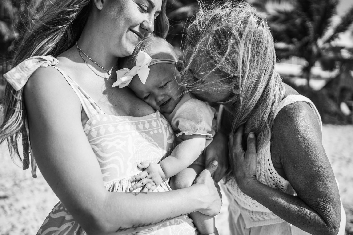 Black and white photo of a mother holding a newborn baby while an older girl leans in to kiss the infant tenderly.