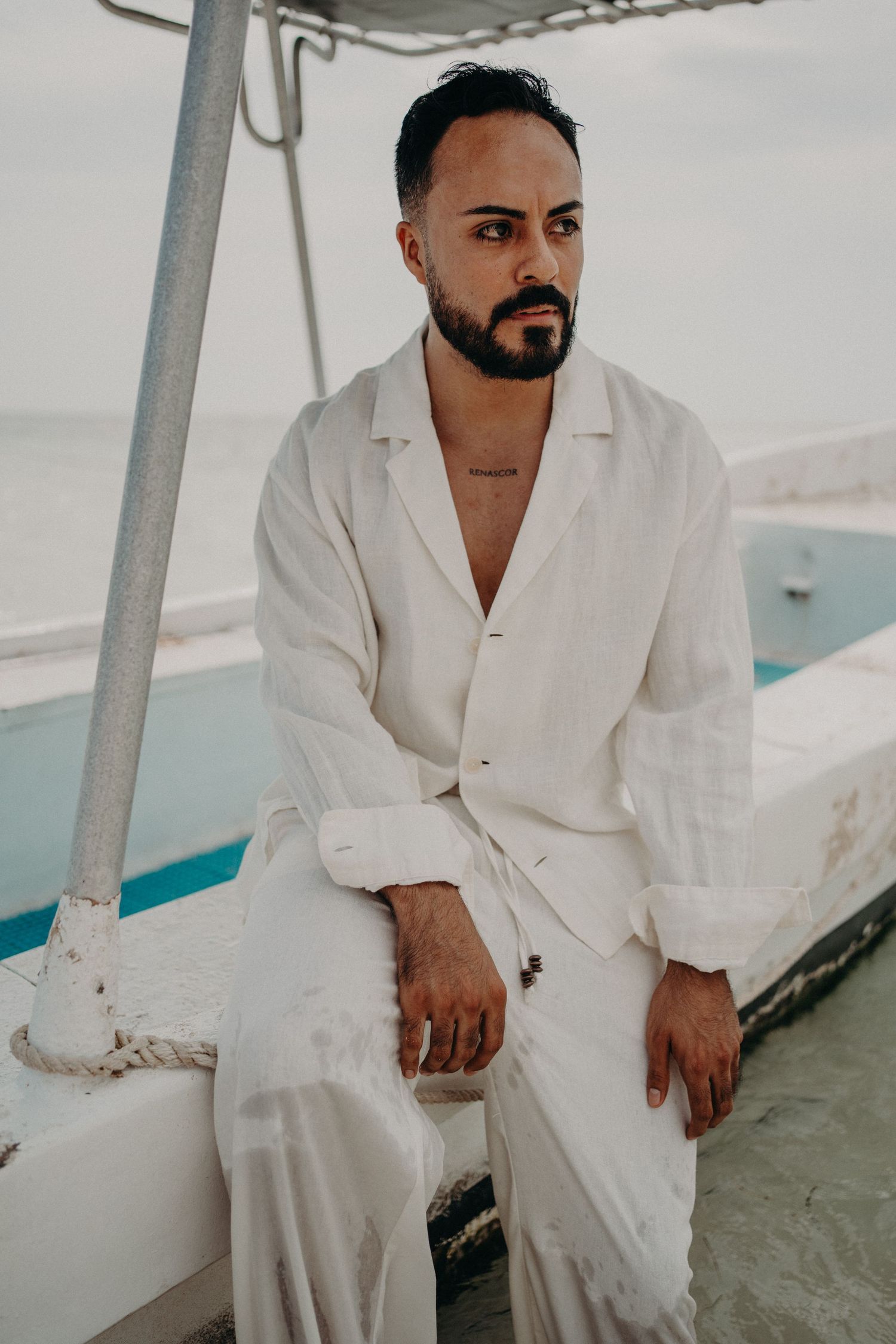 A bearded man in a white linen suit sits on a boat, gazing thoughtfully into the distance near the ocean.