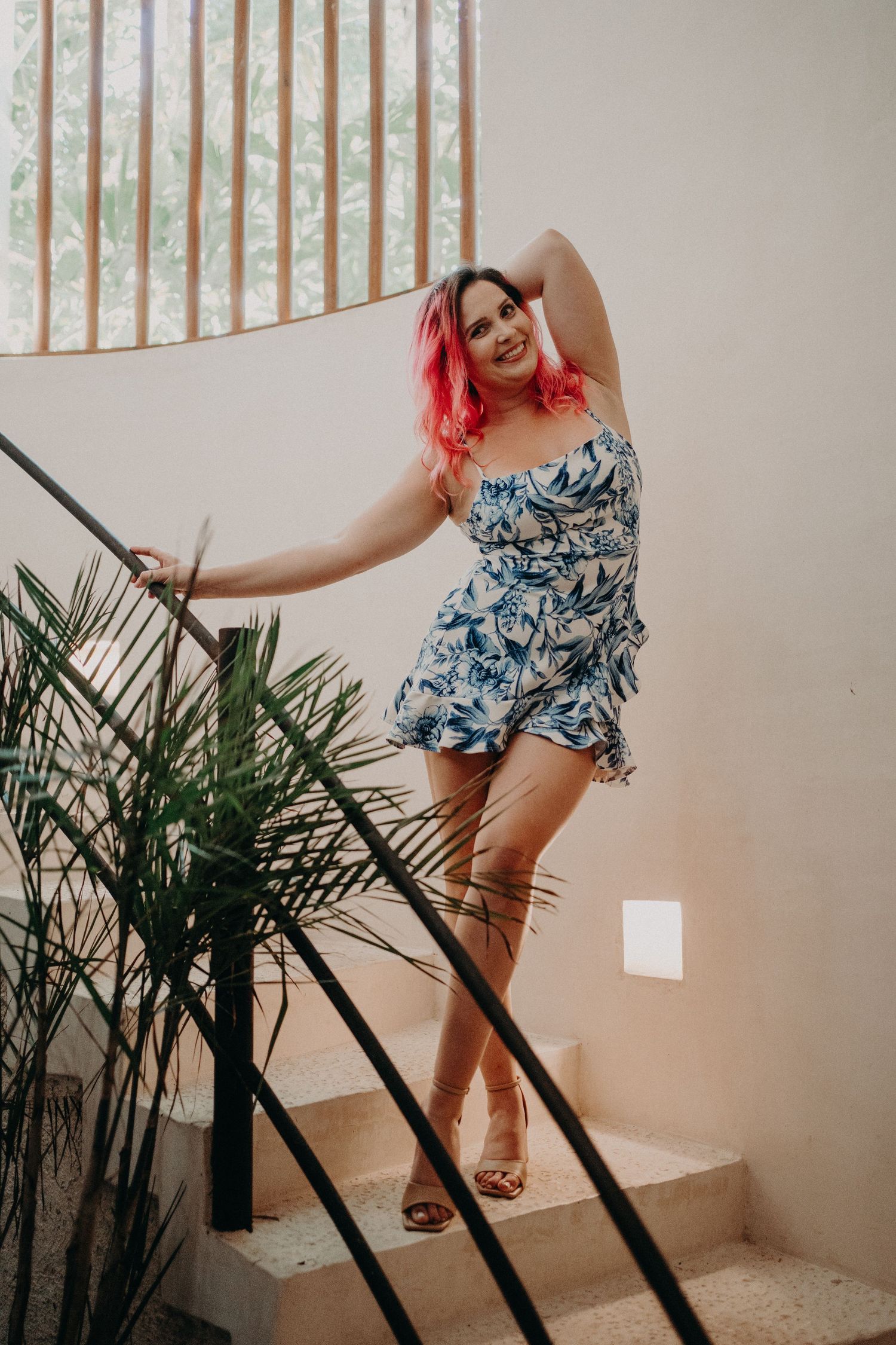 Woman with pink hair in blue floral romper posing on stairs with tropical plants and railing.