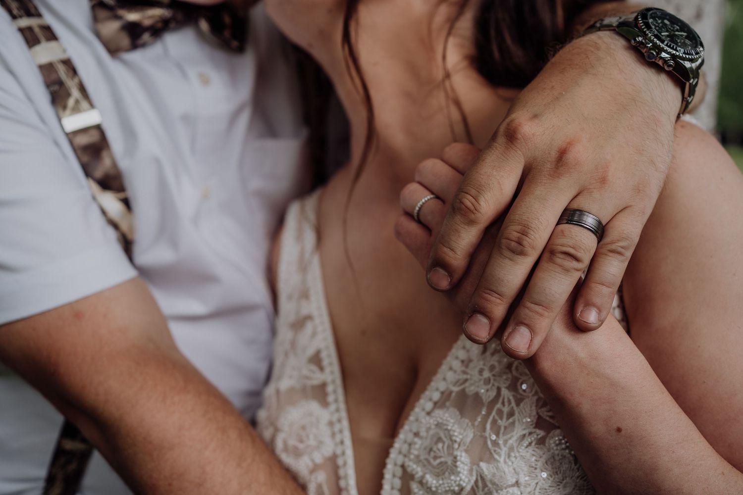 Close-up of newlyweds hands showing matching wedding bands with groom embracing bride in lace dress.