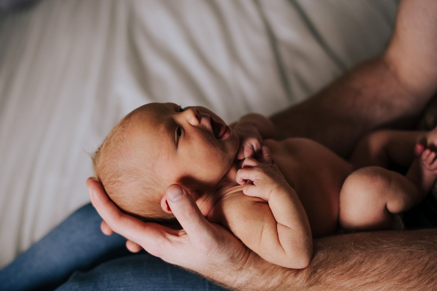 A newborn baby resting peacefully in a parent's gentle hands on a soft white bed.