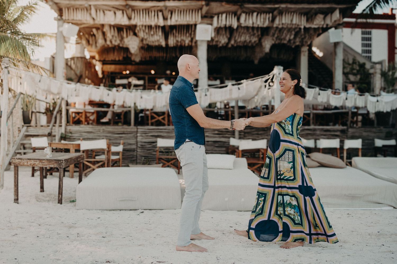 Couple dancing barefoot on white sand beach near thatched-roof restaurant in tropical destination.