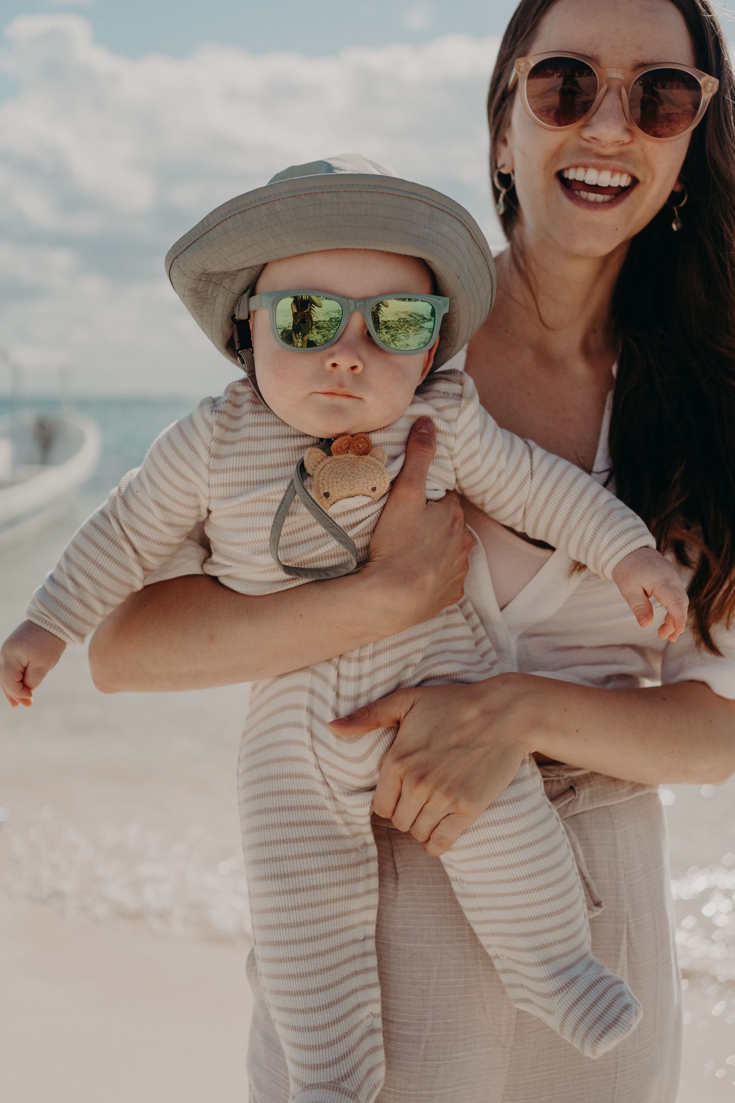 Smiling woman holding a baby wearing sunglasses and a hat on a sunny beach.