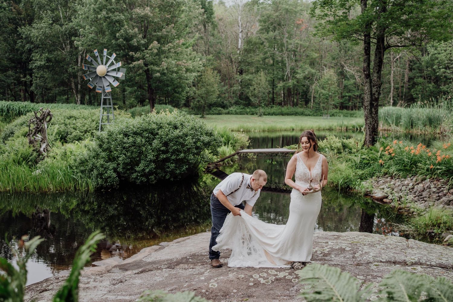 Groom adjusting brides train near scenic pond with windmill and lush greenery in background.