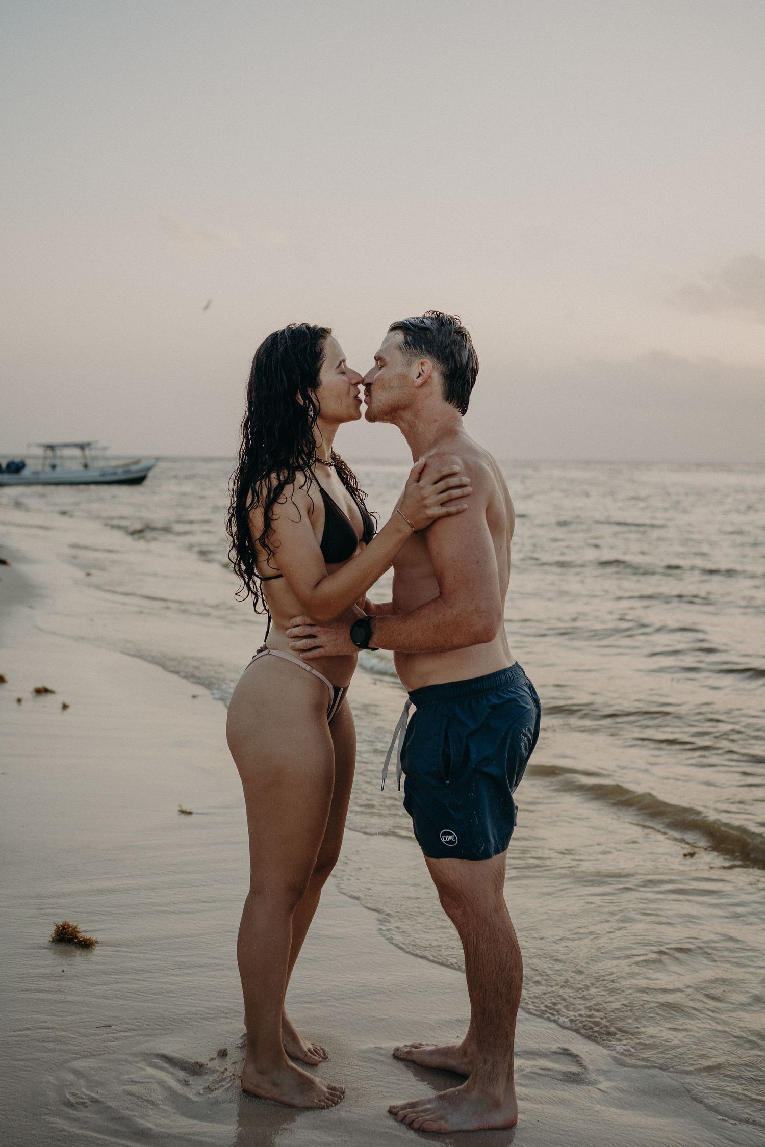 A couple in swimwear shares a kiss while standing in shallow water on a beach at sunset, with a boat anchored in the calm sea behind them.