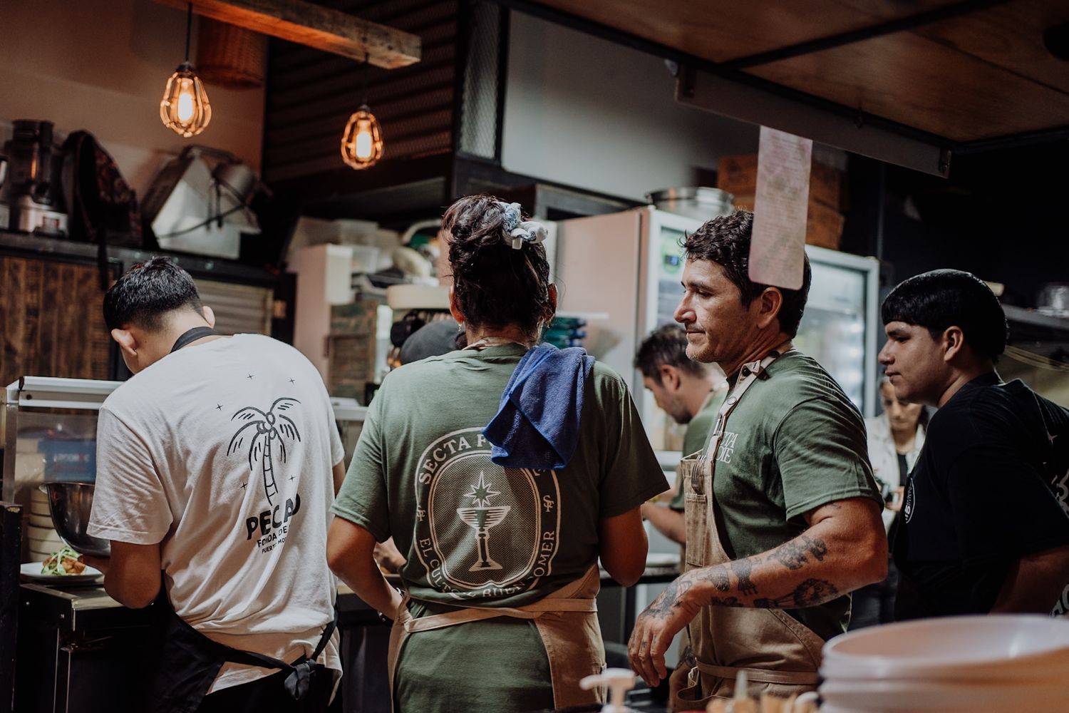 Workers in graphic tees chat inside a busy restaurant kitchen with warm Edison bulb lighting.