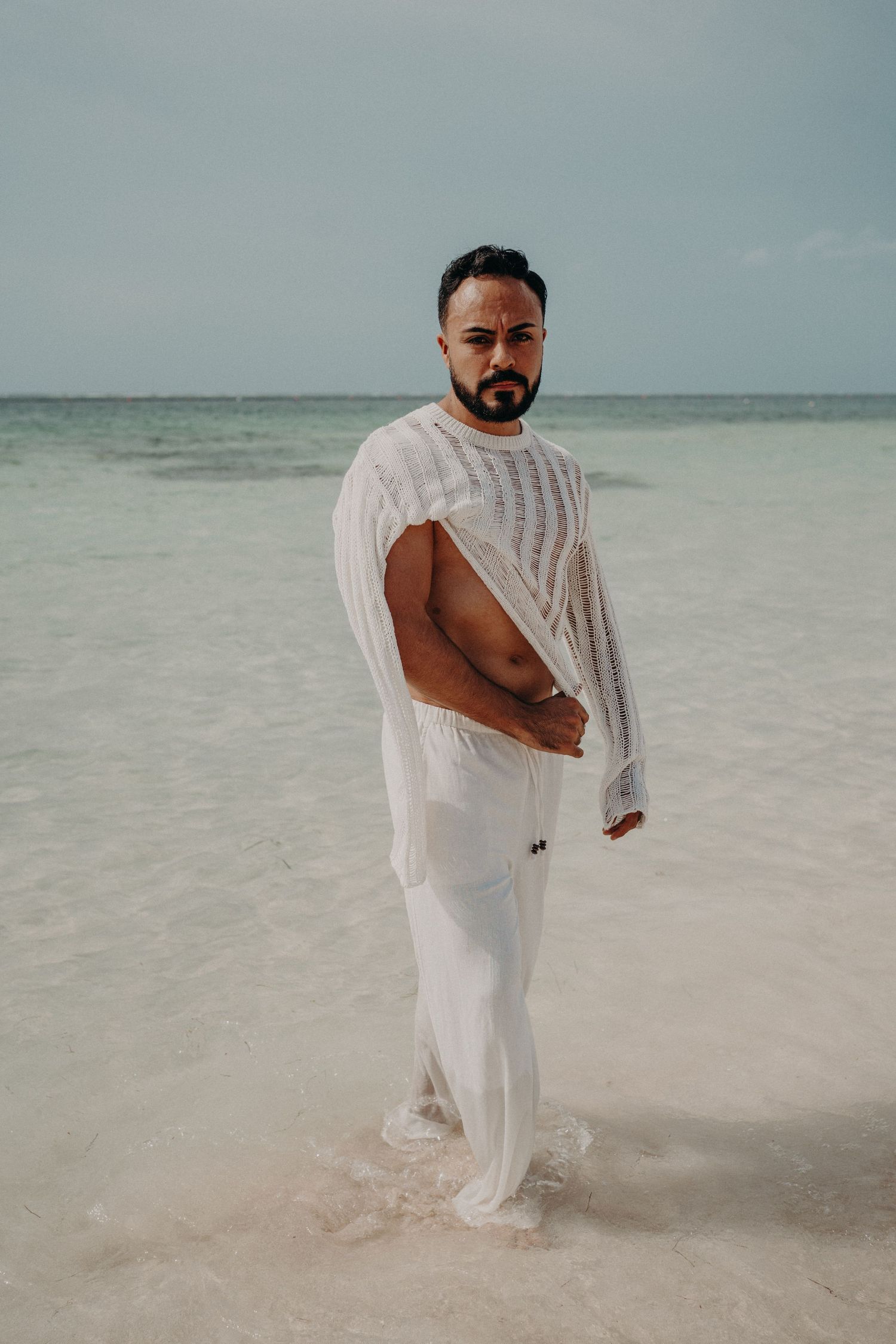 A bearded man in white wet clothing stands in shallow ocean water on a sandy beach.