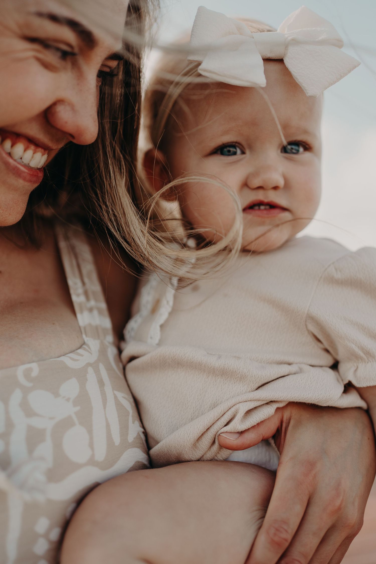 Smiling mother holding her blue-eyed baby girl wearing a white bow headband, both dressed in neutral tones.