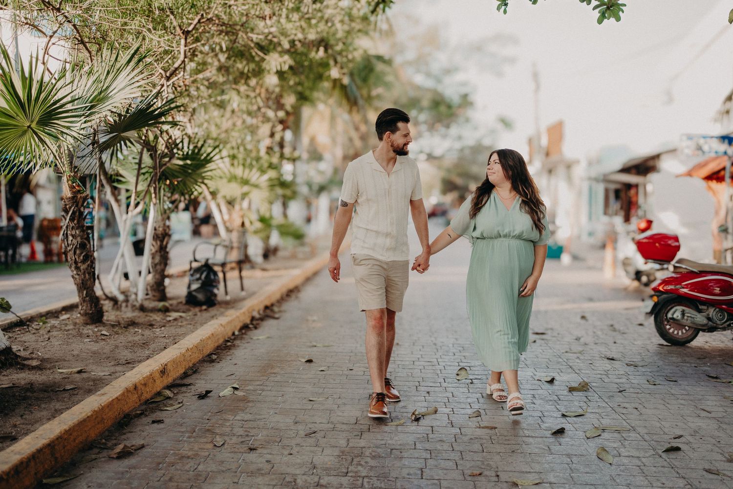 A couple holds hands while walking on a cobblestone street lined with tropical trees and a red scooter in a sunny town.