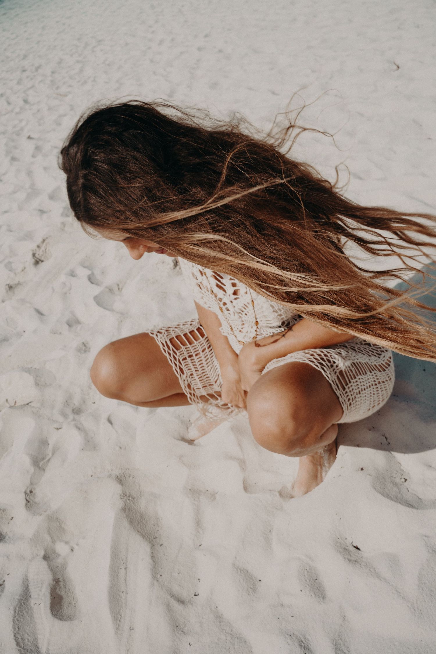 Woman with long wavy hair crouching on white sand in a crochet cover-up, with wind blowing her hair across her face.