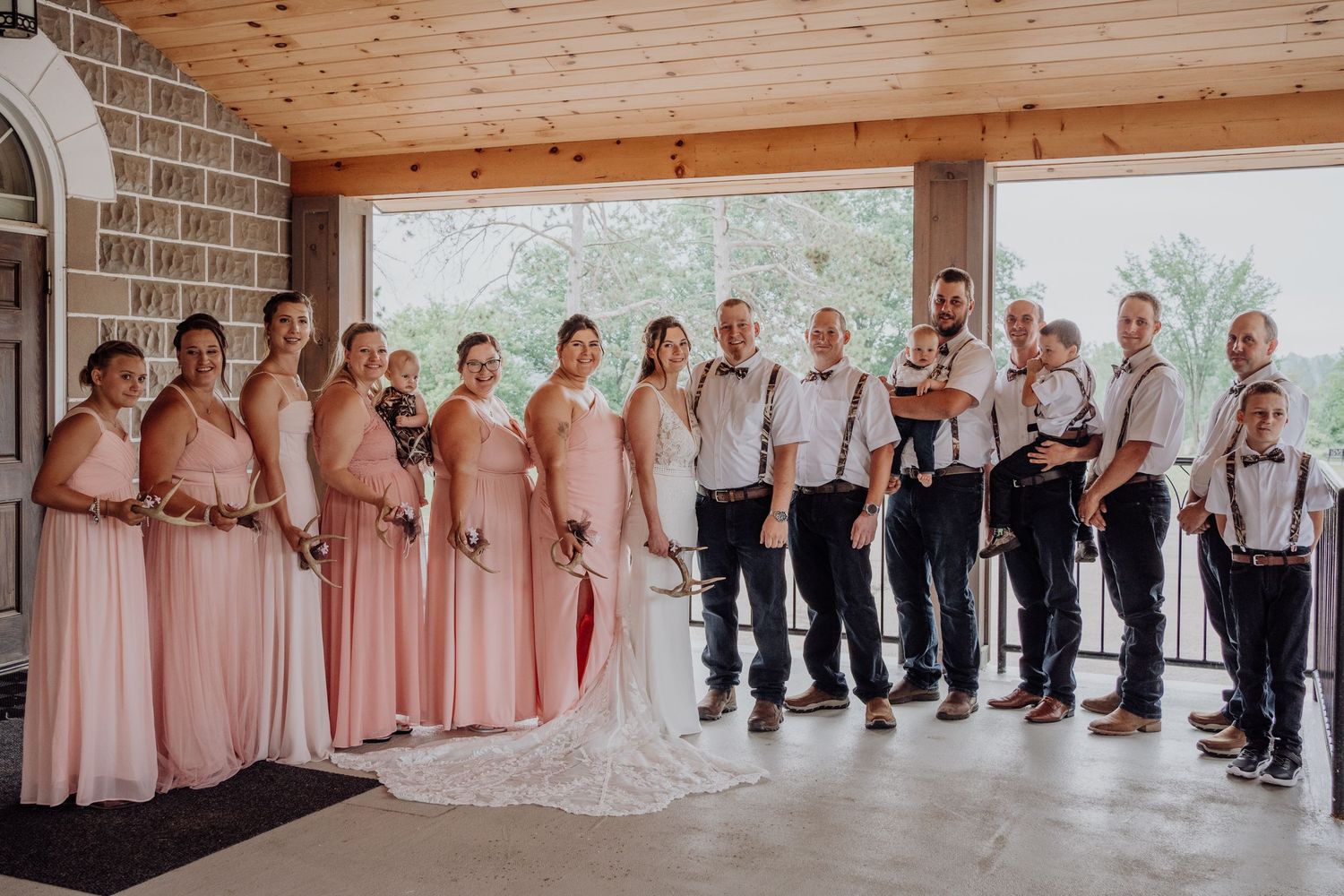 Full wedding party in blush pink and navy attire poses together under an outdoor pavilion.