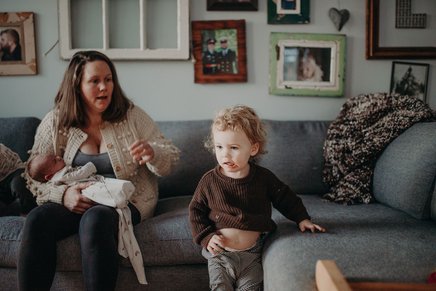 A mother sits on a gray sofa holding a newborn baby while a toddler stands beside her in a cozy living room.