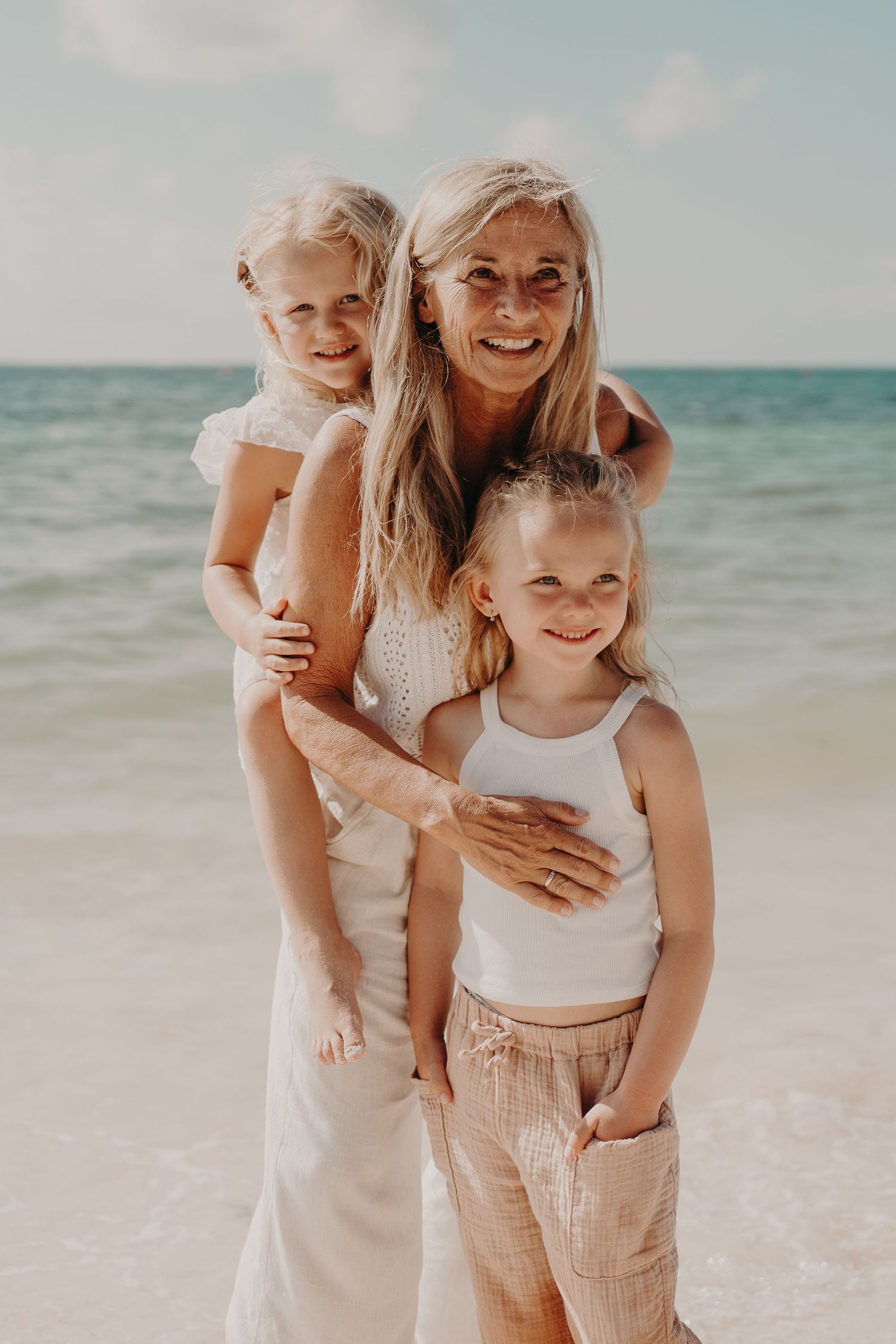 A smiling grandmother poses on a beach with two young blonde girls, all dressed in white and beige outfits.