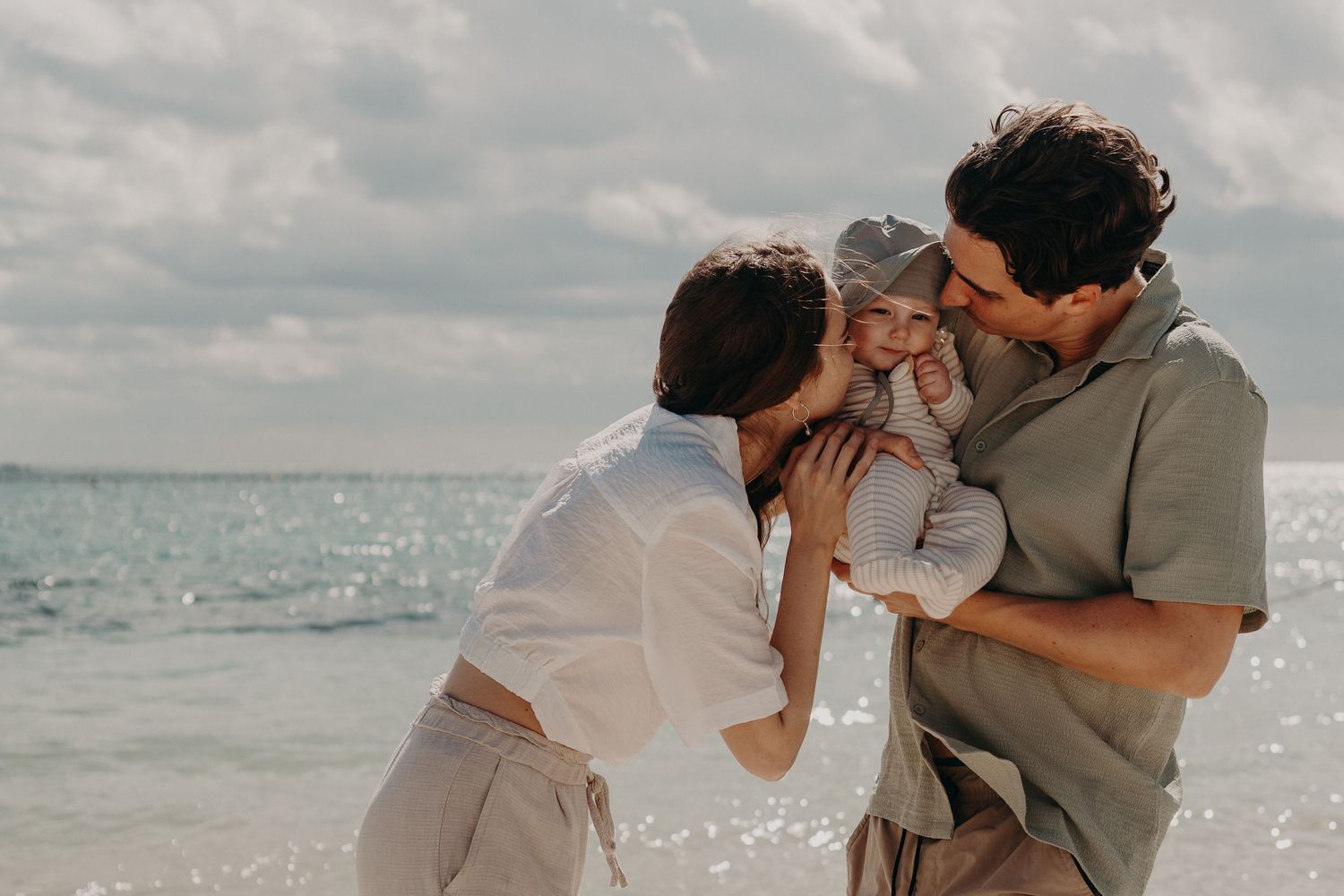 A happy family with a baby enjoys a beach day, with parents lovingly kissing their child by the ocean.