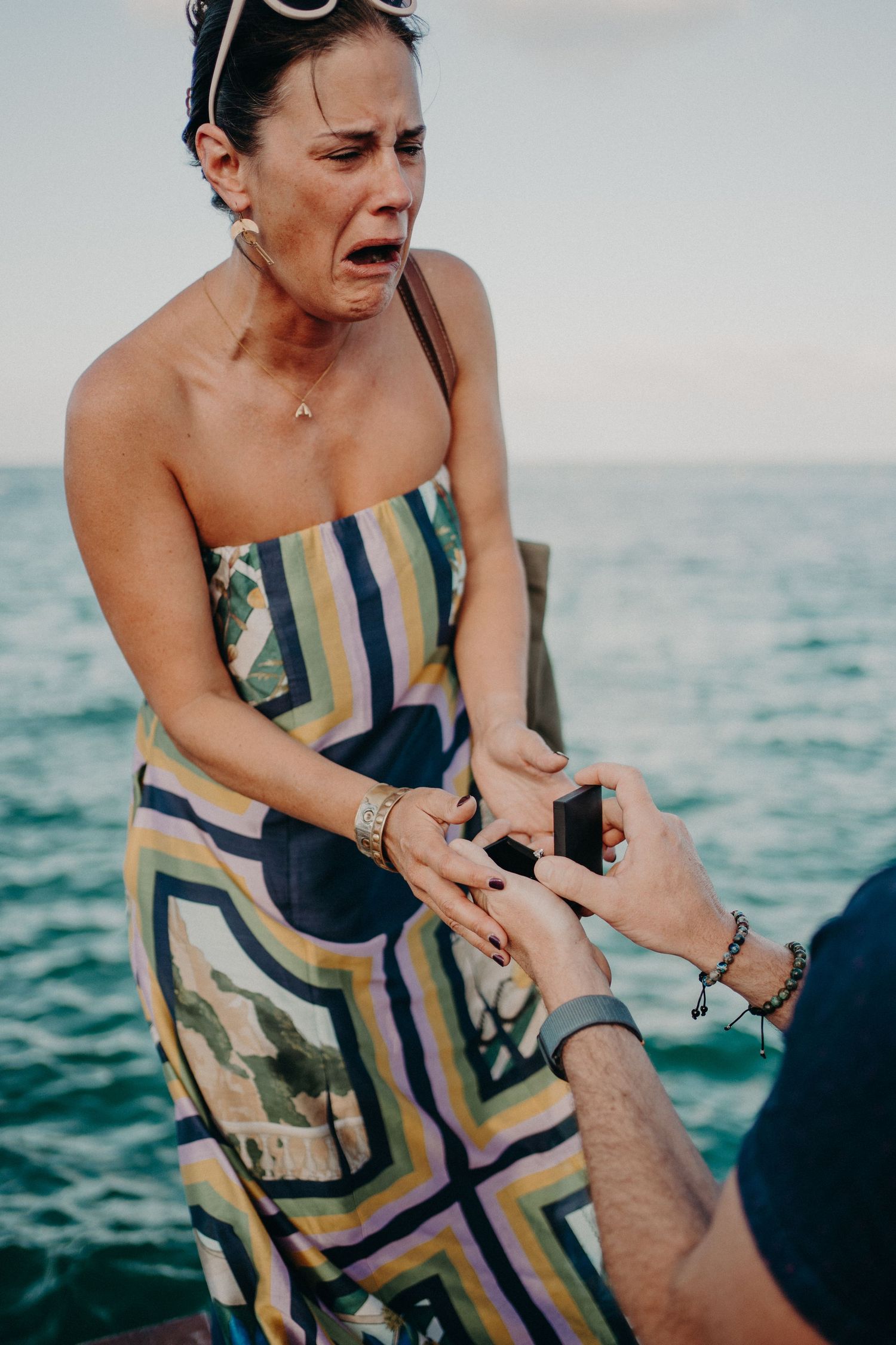Woman in striped strapless dress crying with surprise as man places ring on her finger by the ocean.