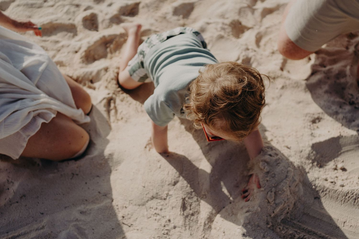 Young child with curly hair playing in sand at the beach, digging with hands while adults sit nearby.
