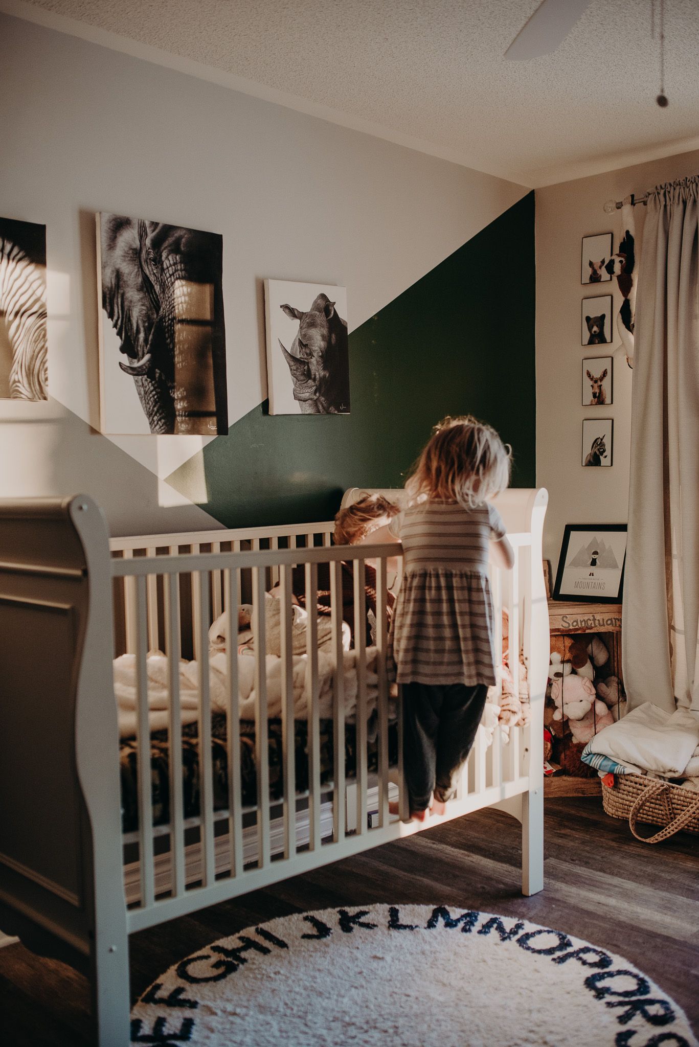 A toddler stands beside a white crib in a safari-themed nursery with green accent wall.