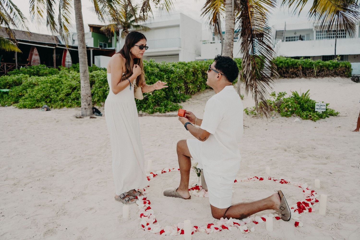 Man in white kneeling on one knee proposing to a smiling woman in a long white dress on a tropical beach inside a heart of red rose petals.
