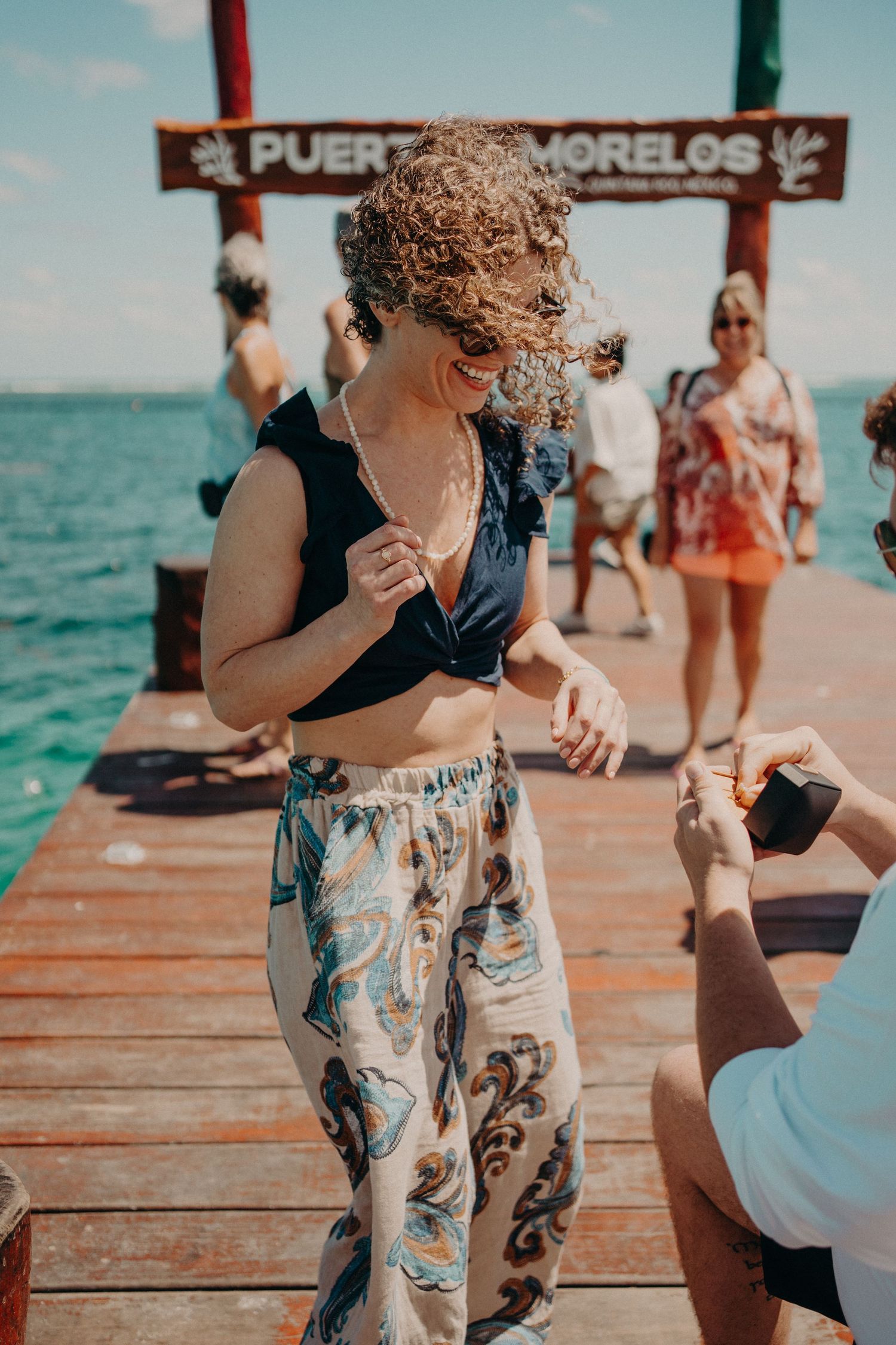 Woman in crop top and paisley pants on a wooden pier in Puerto Morelos, Mexico, receiving a ring box proposal.