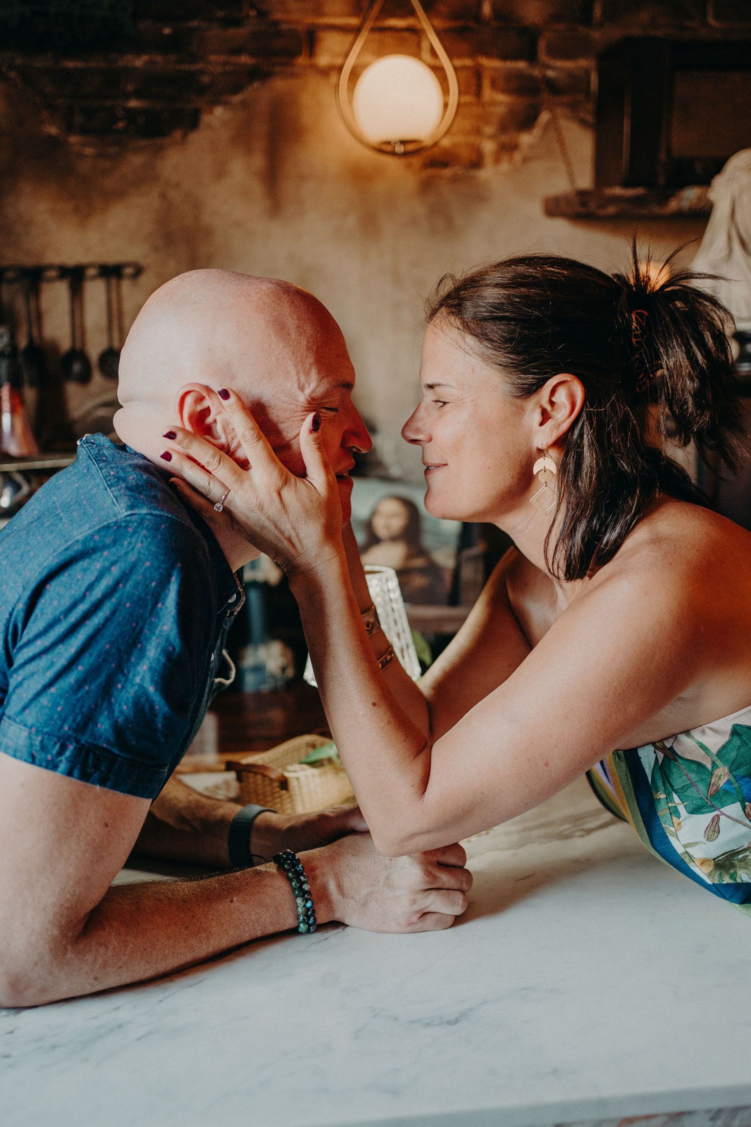 A couple shares a tender moment, woman cradling man's bald head, leaning in for a kiss at a marble counter.