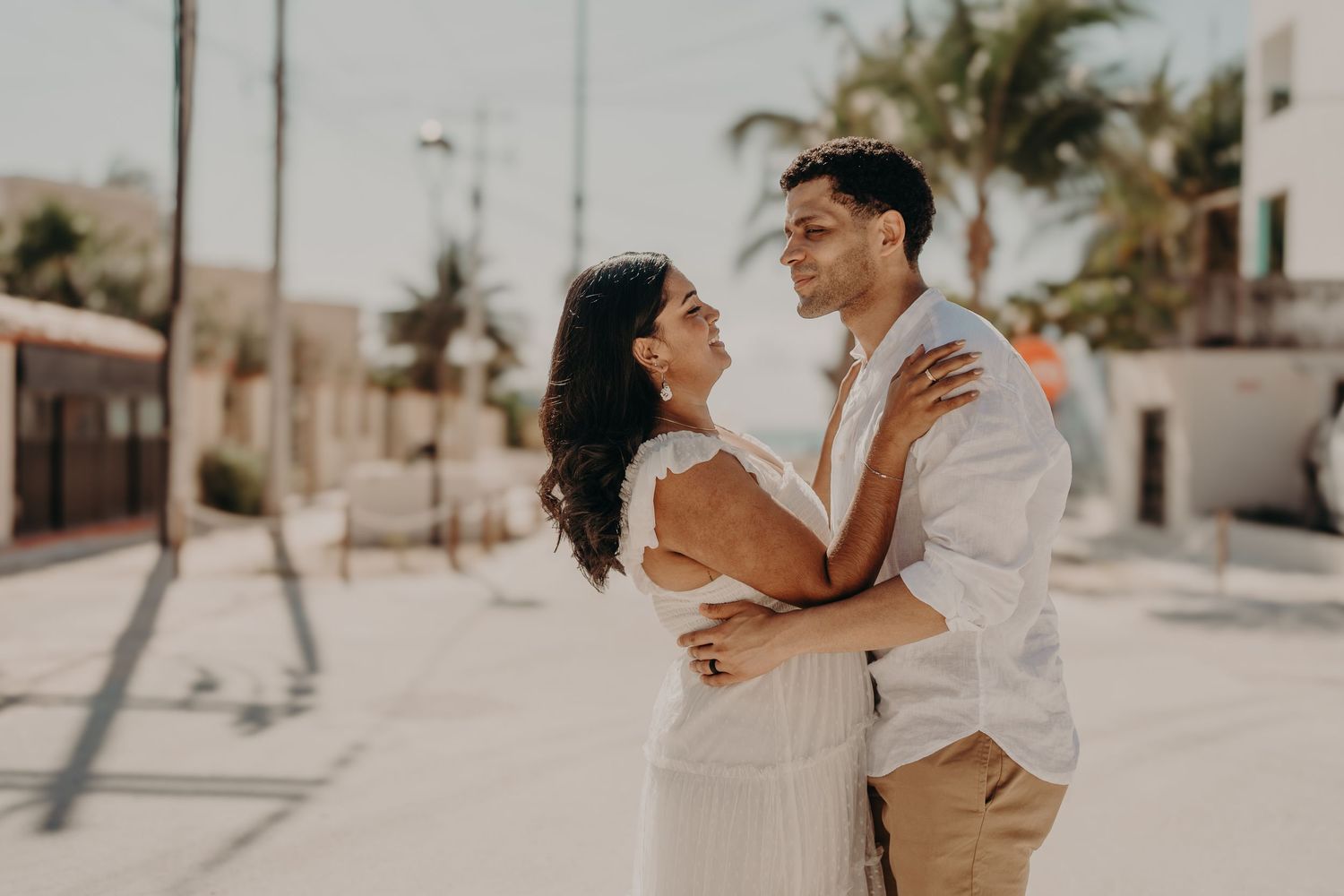 A couple embraces and gazes at each other outdoors in a sunny coastal town, dressed in white and beige.
