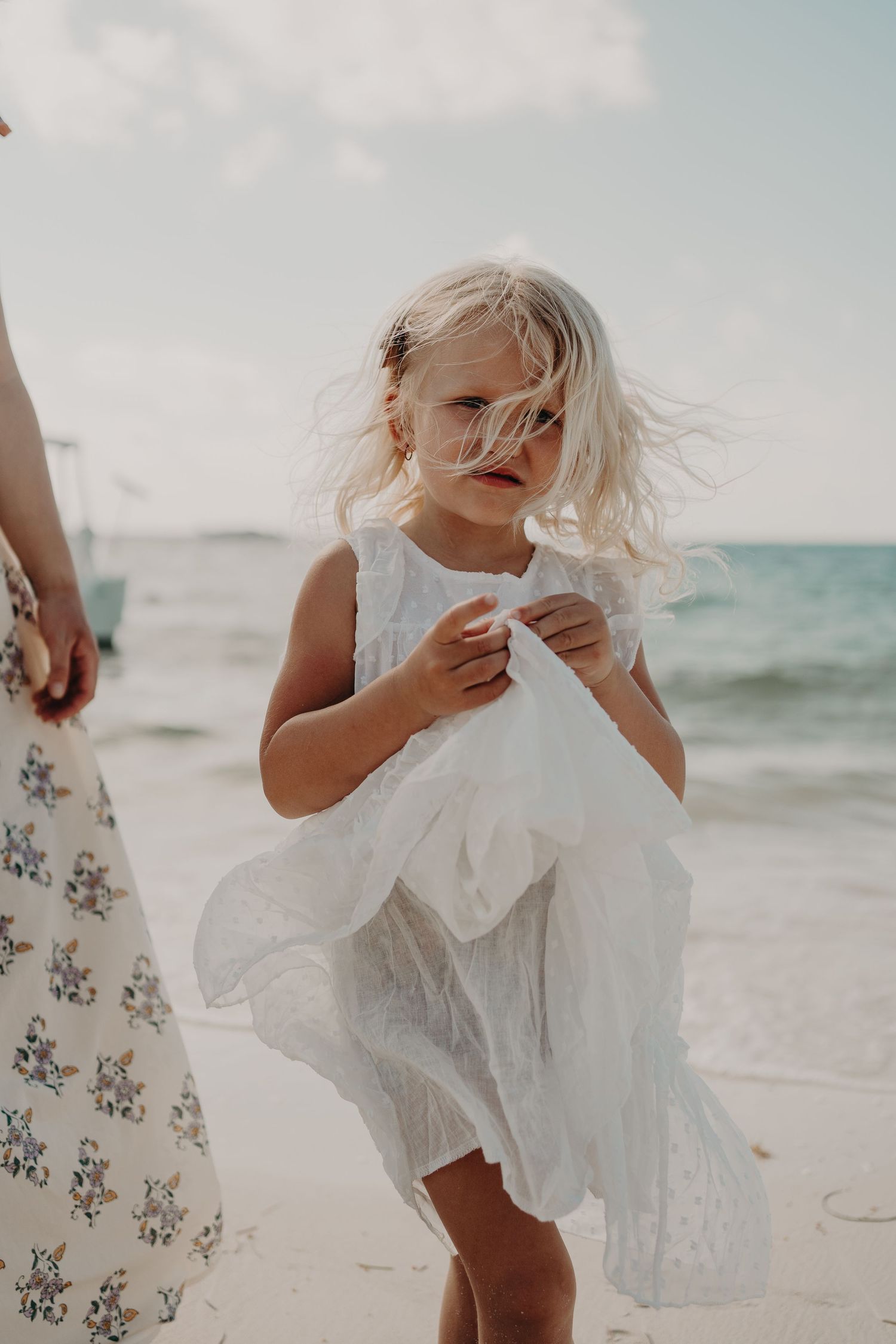 Young blonde girl in white dress playing on sandy beach with ocean waves in background on cloudy day.
