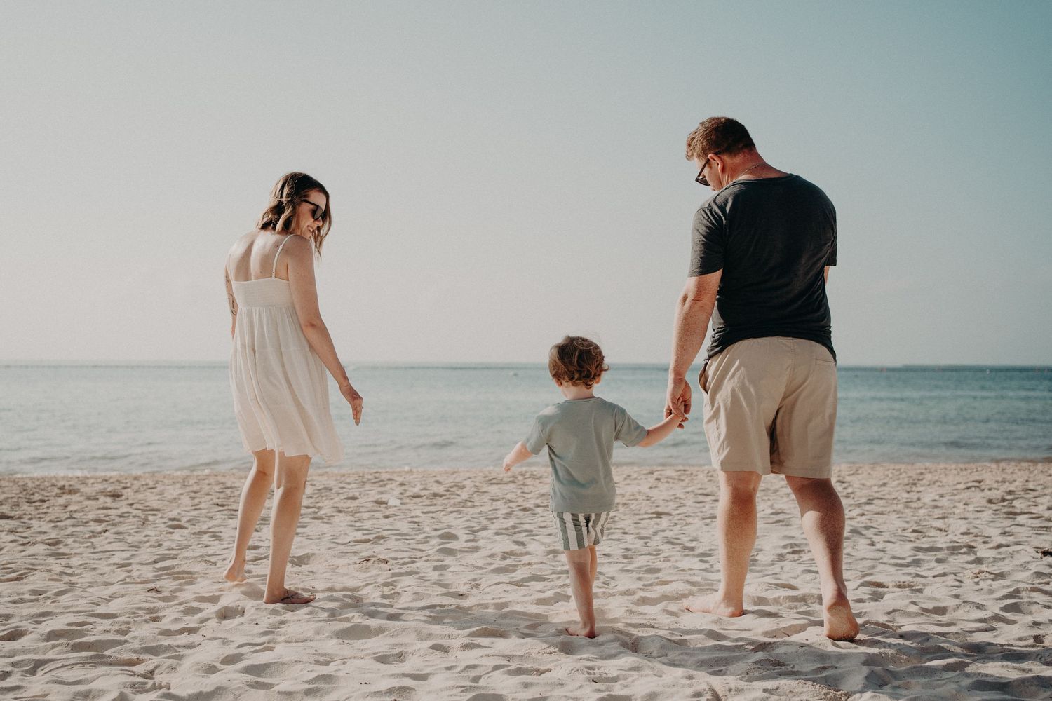 Family of three holding hands on a sandy beach, parents and toddler walking together by the ocean on a sunny day.