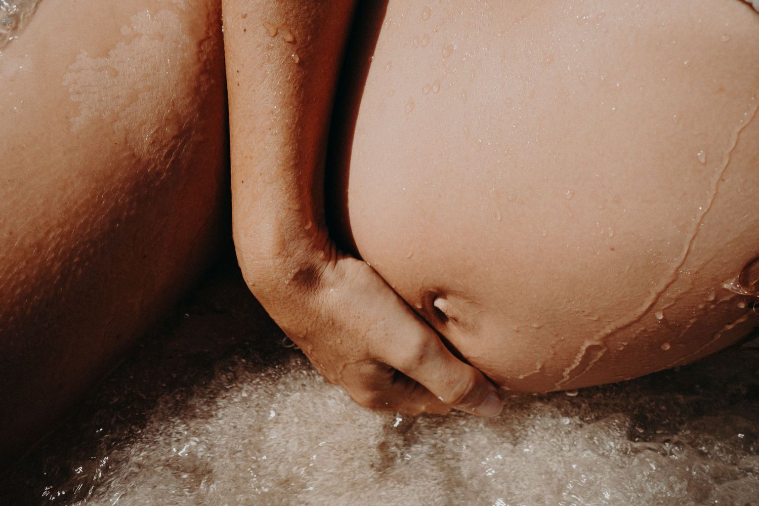 Close-up of a hand resting on a pregnant belly with water droplets and sparkling foam in warm, sunlit maternity photography.