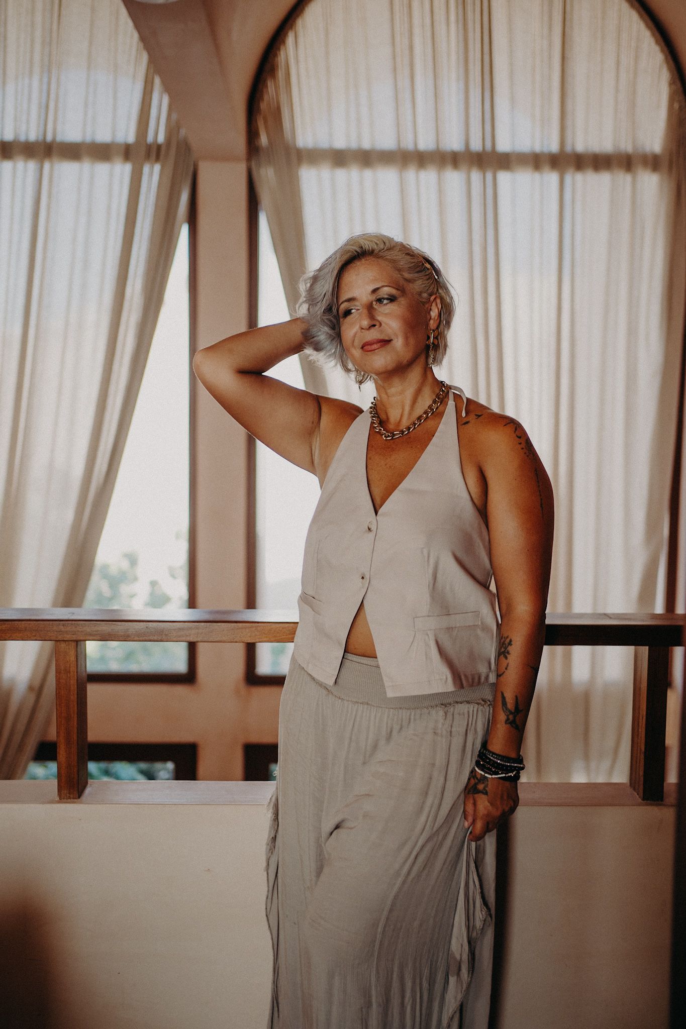 Elegant older woman with gray hair poses in a beige vest and maxi skirt, wearing a necklace, on a balcony.