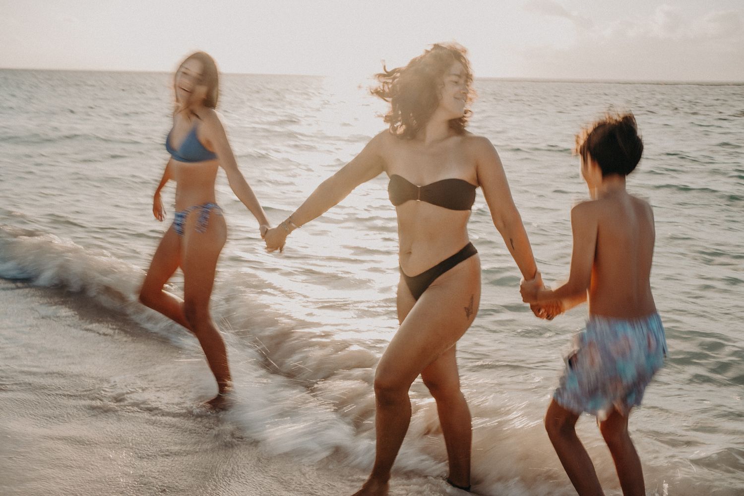 Three women in swimsuits holding hands and playing in ocean waves on a sandy beach at sunset.