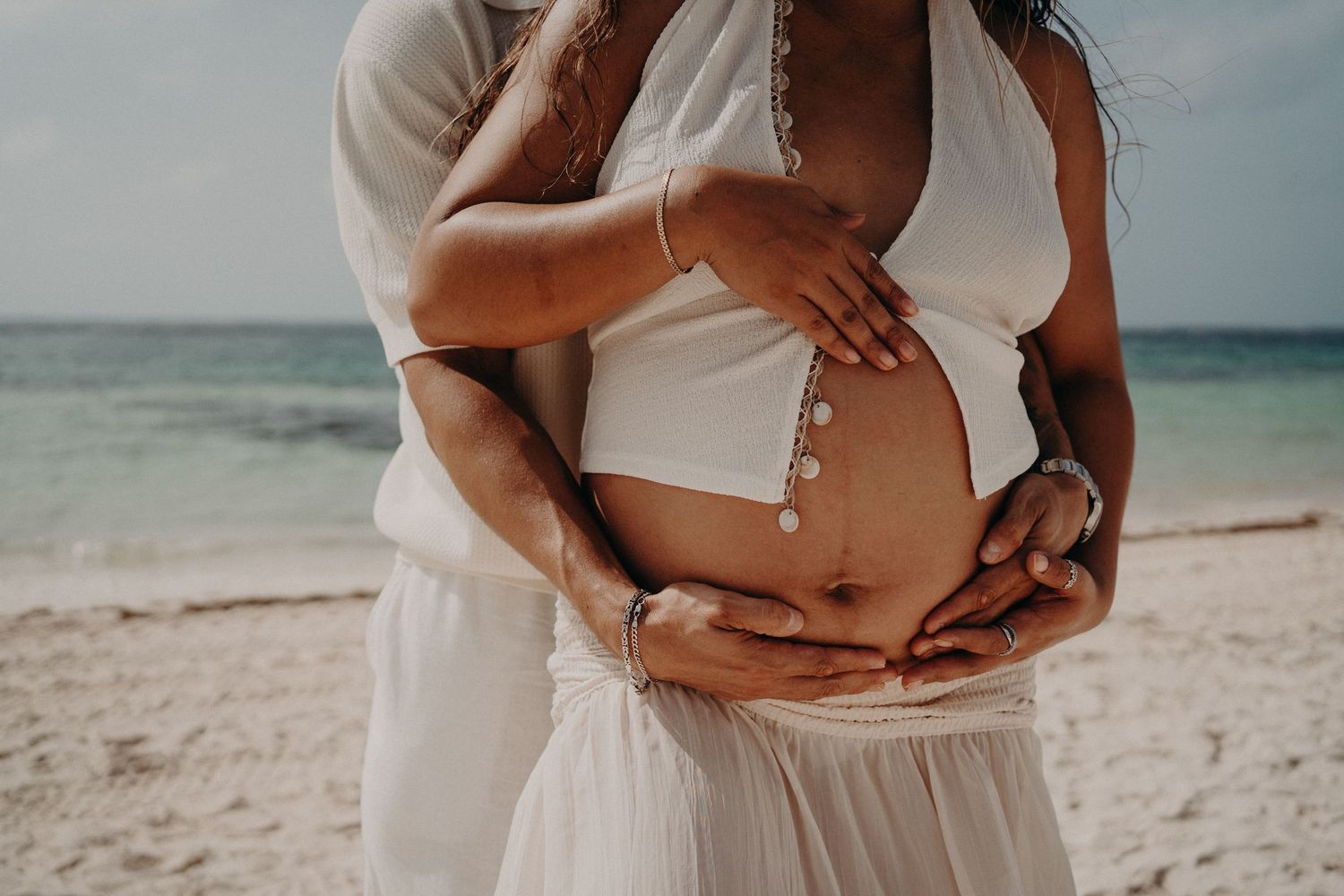 Couple embracing on a beach, hands cradling a pregnant belly, wearing white outfits with ocean in the background.
