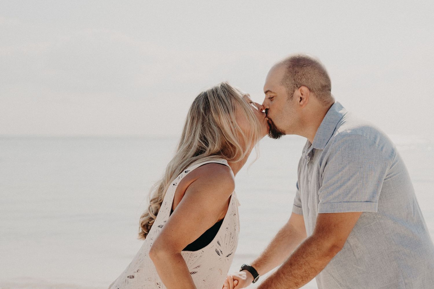 Couple sharing a romantic kiss on the beach with calm ocean waters in the soft background.