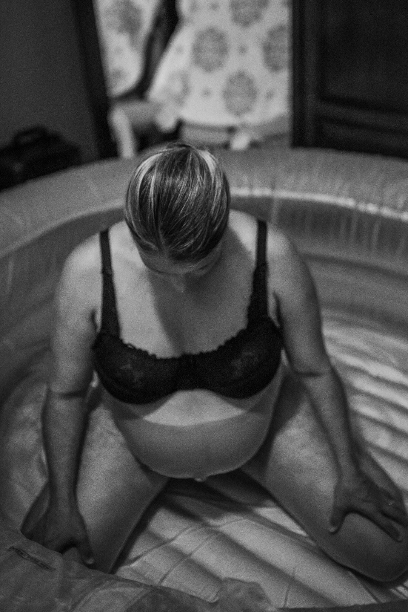 Black and white photo of a pregnant woman in a bra kneeling in an inflatable birthing pool.